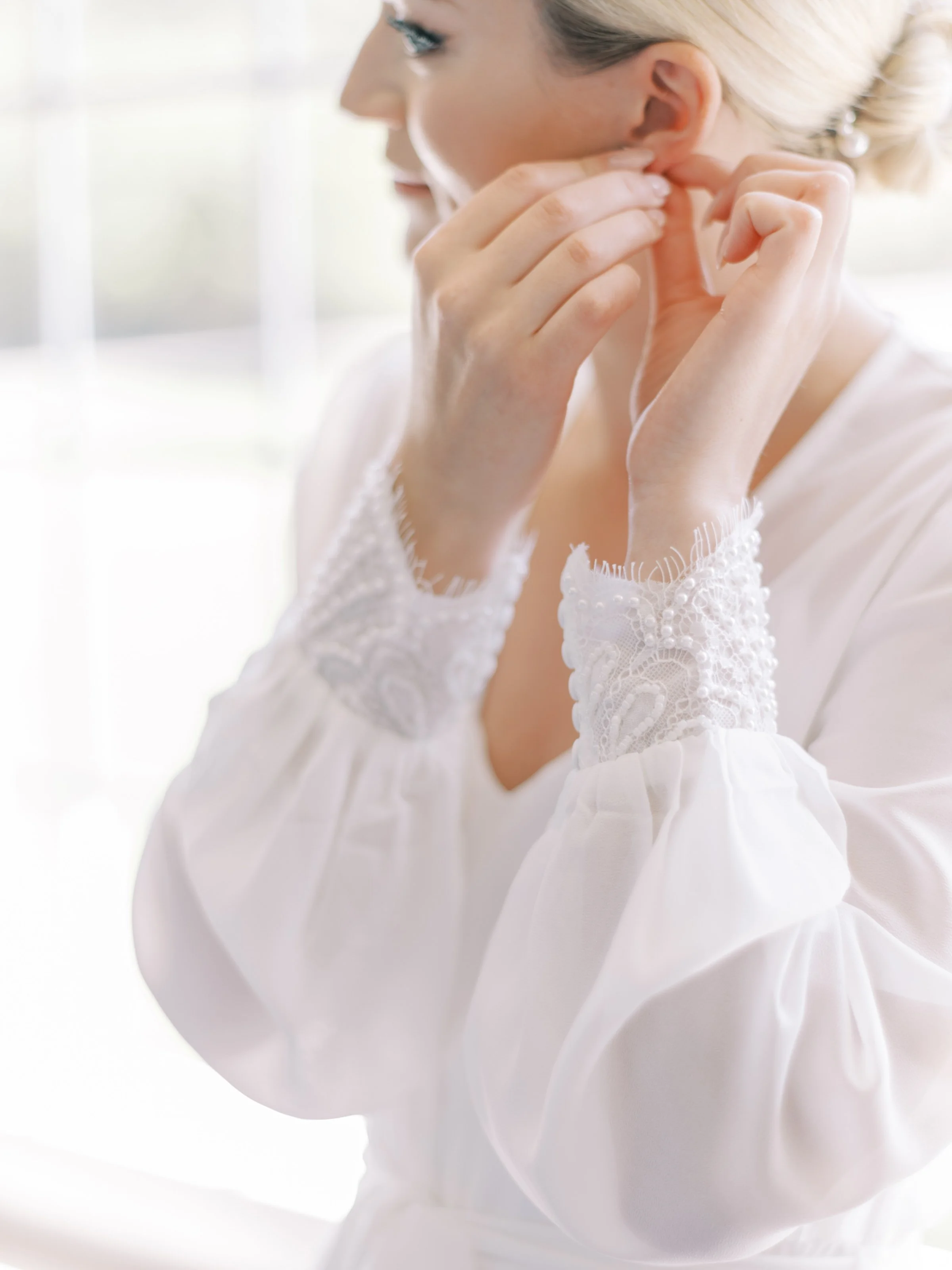 A woman in a white top with lace details adjusting her earrings in front of a bright window.