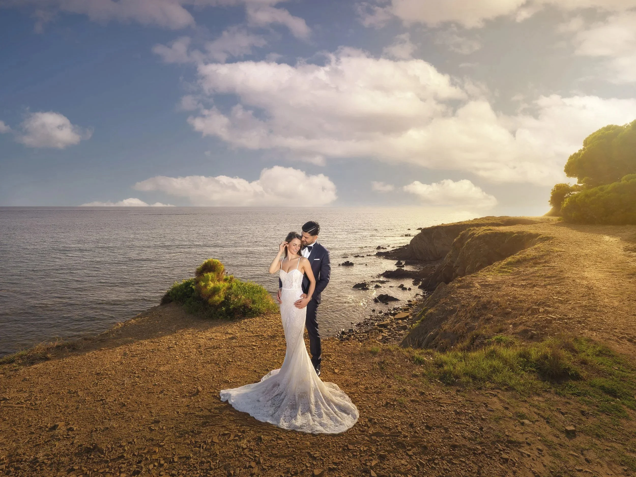 A couple dressed in wedding attire standing on a rocky coastline with the ocean and a partly cloudy sky in the background. The bride is wearing a white gown and the groom a dark suit. The sun is setting or rising, casting a warm glow.
