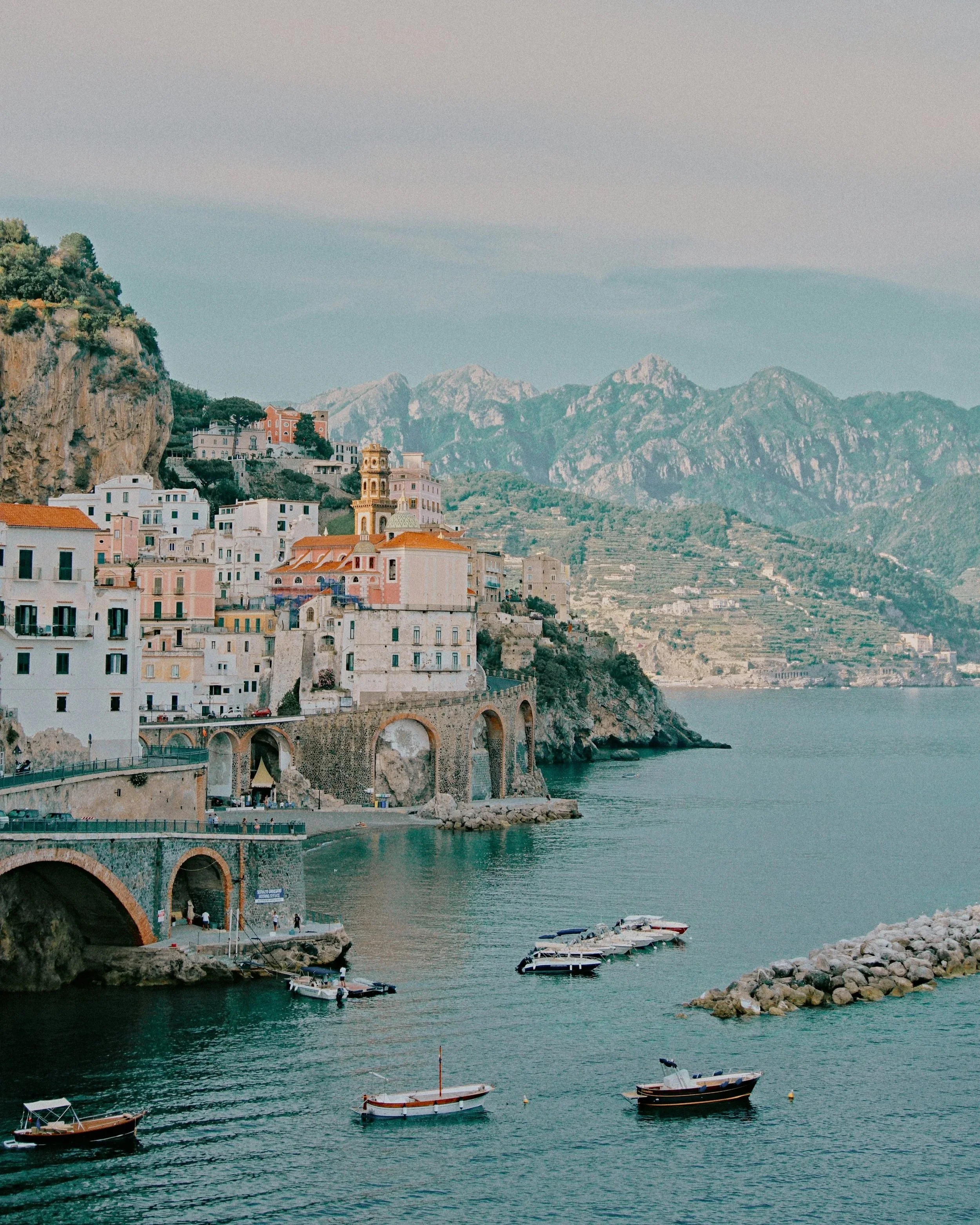 Coastal town with colorful buildings along a cliffside, mountains in the background, boats in the water, and a walkway with people.