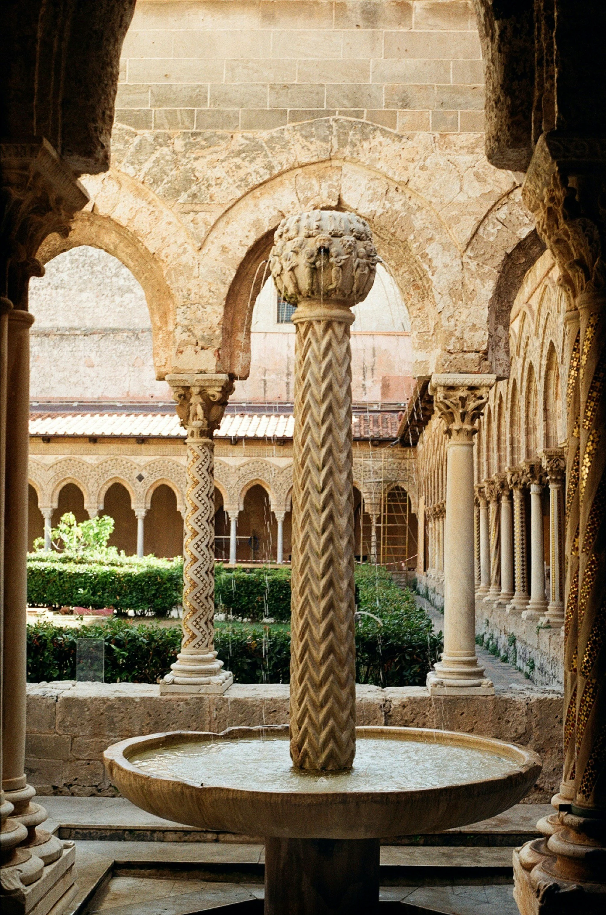 Ancient stone fountain with water flowing from carved sculptures of lions, situated in a cloister with arches and columns, overlooking a garden.