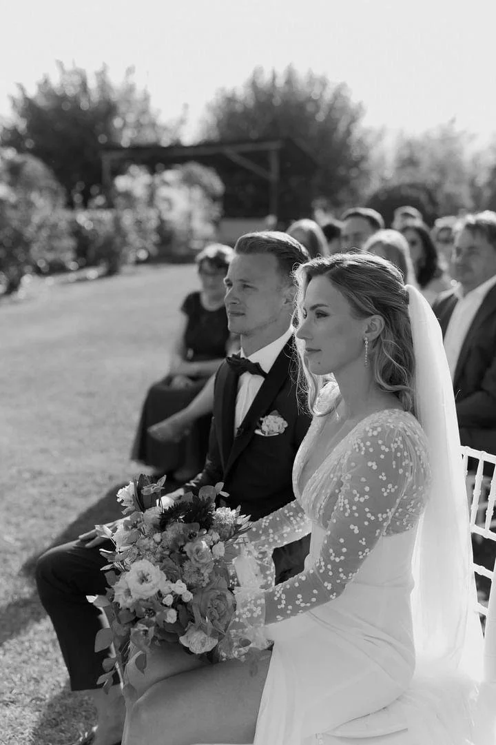Black and white photo of a bride and groom seated outdoors during a wedding ceremony, with guests in the background.