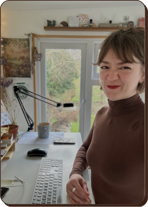 EJ has short brown hair and a brown turtleneck taking a selfie in a home office. Behind her is her white desk layout, a beautiful little shelfie with natural treasures on it like rocks and shells with greenery in the background through the windows.