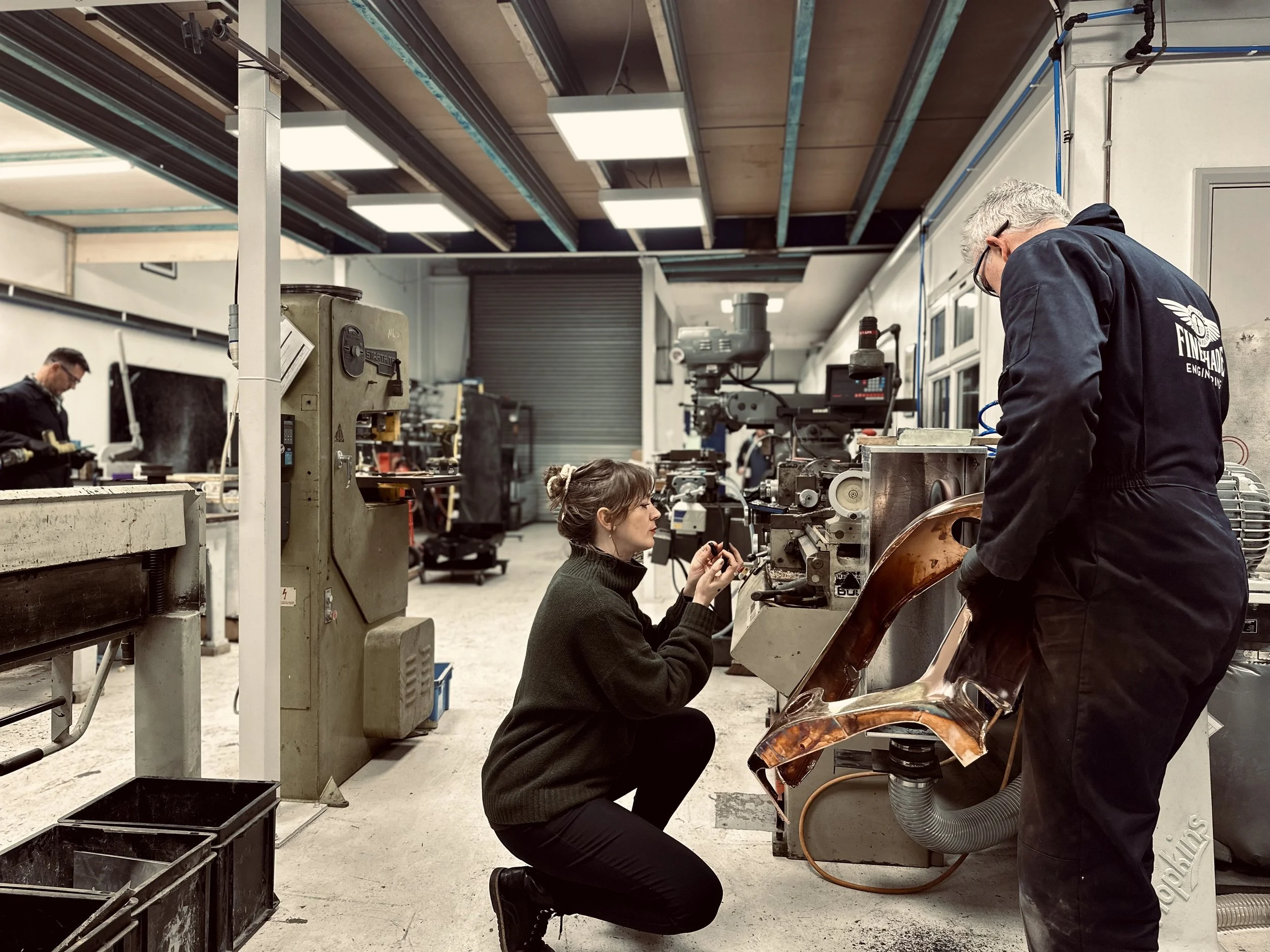 EJ is kneeling in Fineshade Engineering's workshop filming Ben operating a large industrial machine polishing a vintage car radiator shell for their Instagram account.