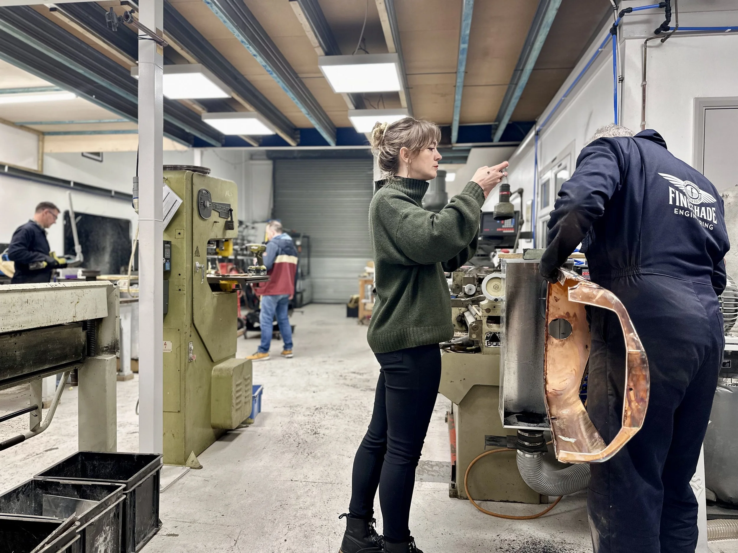 EJ is working with a man in a dark blue work overalls labeled 'Fineshade Engineering' on a copper vintage car radiator on industrial equipment in a workshop. Two other workers are visible in the background operating machines.