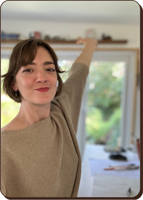 EJ has short brown hair and a beige top smiling and reaching up to a shelf in a bright wendy office with greenery outside.