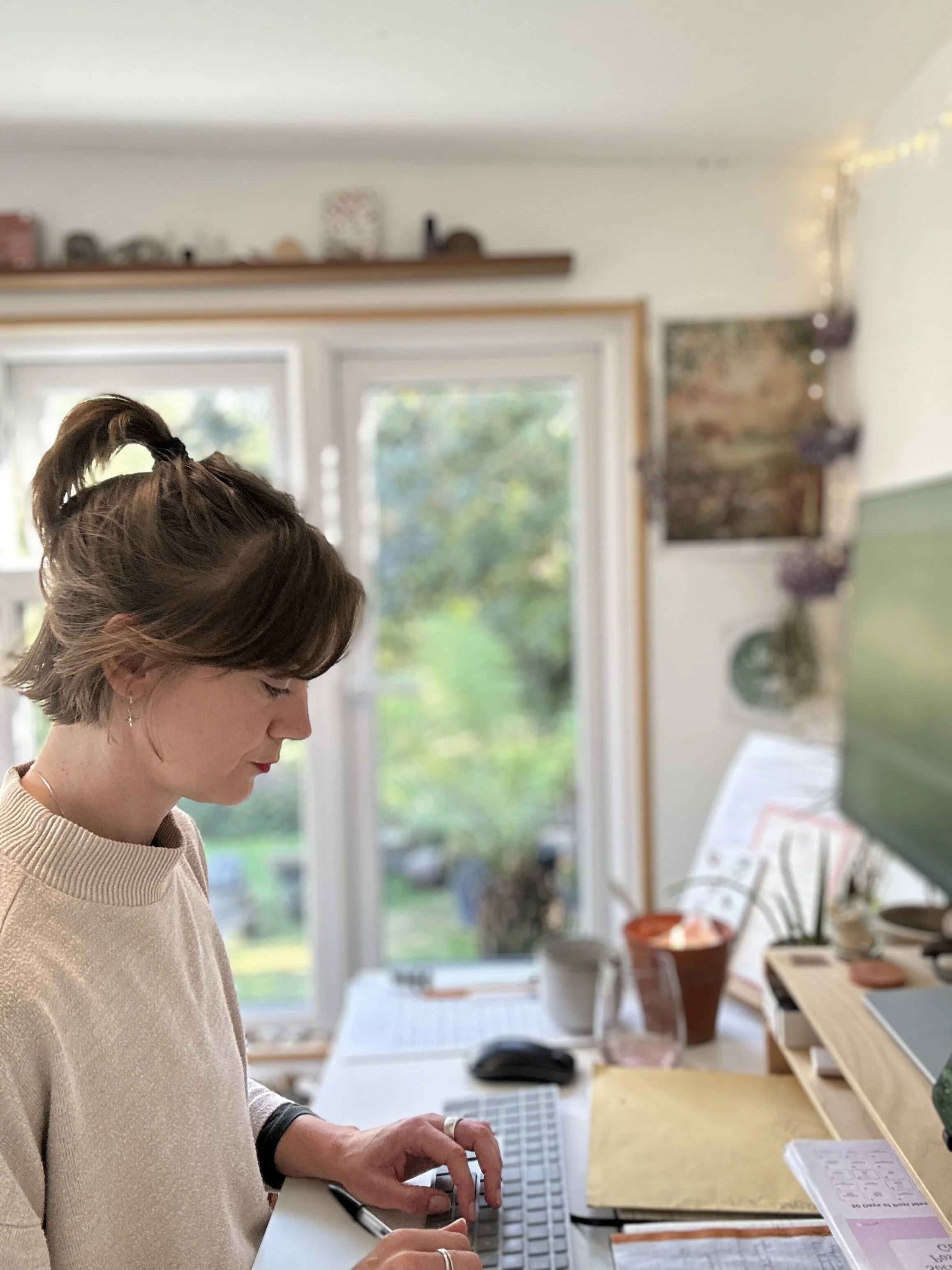 EJ has short brown hair and a beige sweater and is head down working at her desk in her home wendy office in the garden, with a large window showing greenery outside.