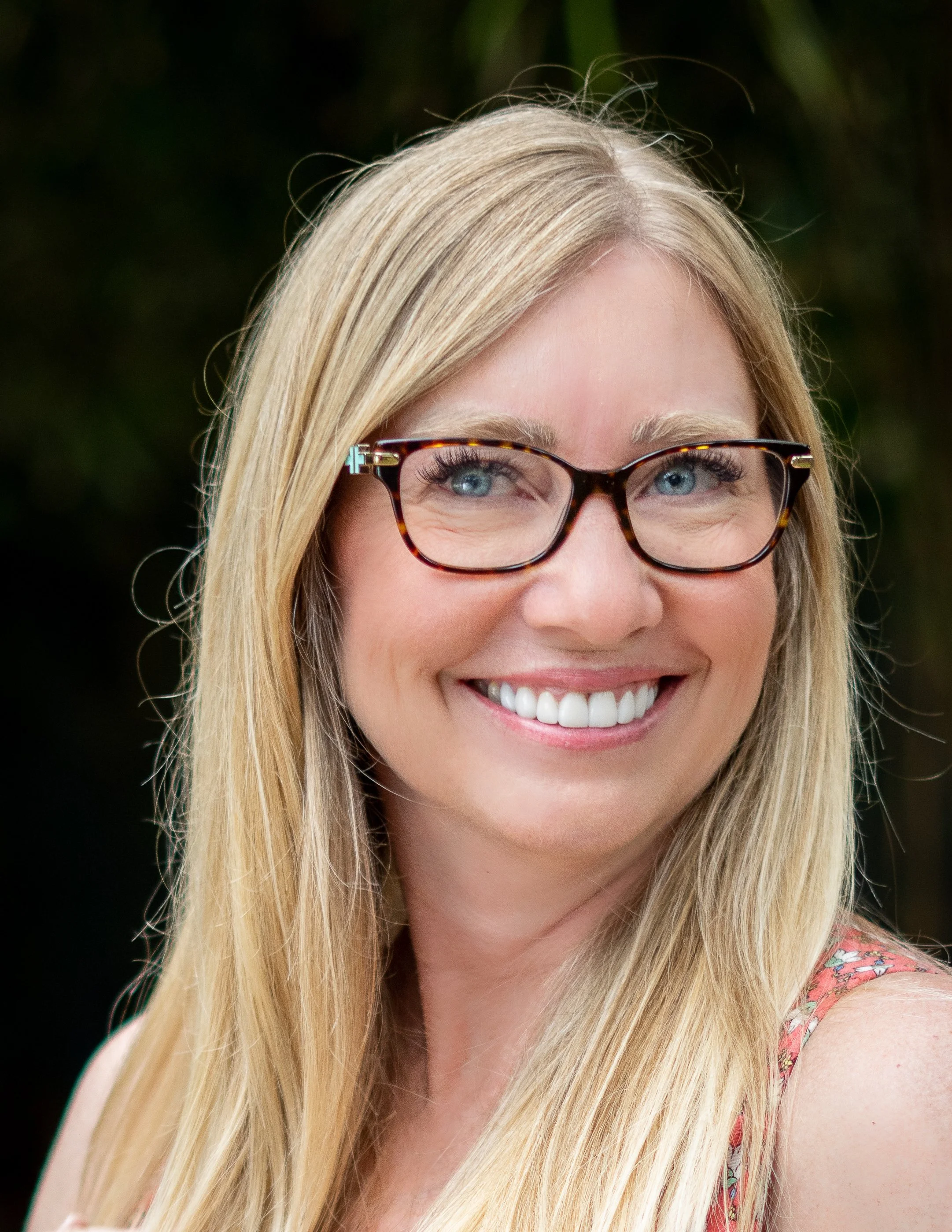 A smiling woman with long blonde hair, wearing tortoiseshell glasses and a floral top, outdoors with blurred greenery in the background.
