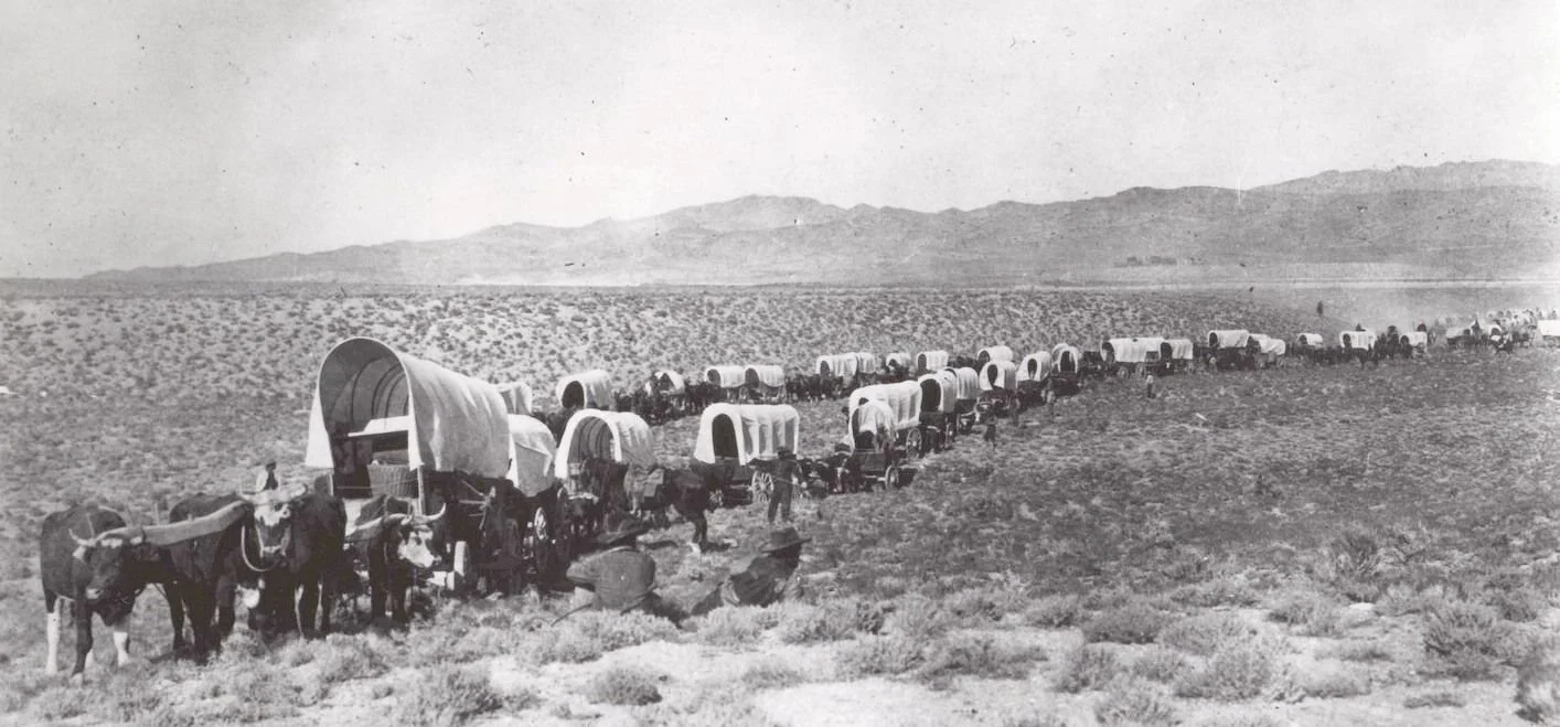 A long line of covered wagons with oxen in a desert landscape, with mountains in the background.