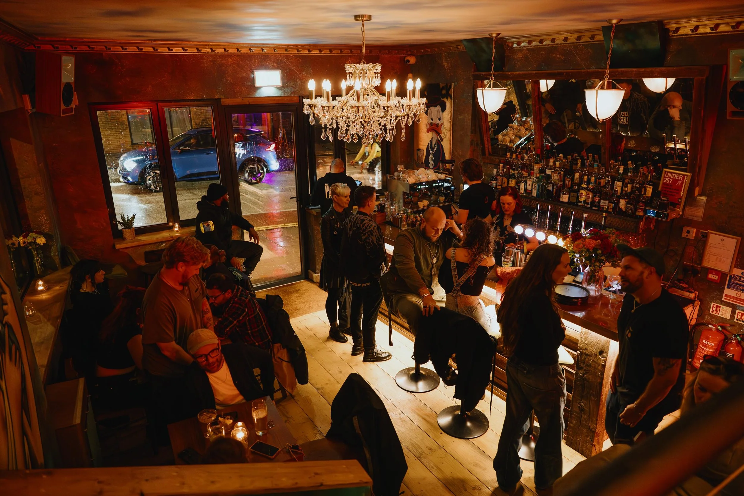 Interior of a lively bar or pub with people socializing, a bartender behind the counter, a chandelier hanging from the ceiling, and large window showing cars on the street outside during nighttime.