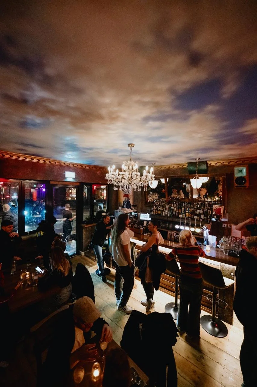 Interior of a bar with people socializing, a chandelier hanging from the ceiling, and a cloudy sky painted on the ceiling.
