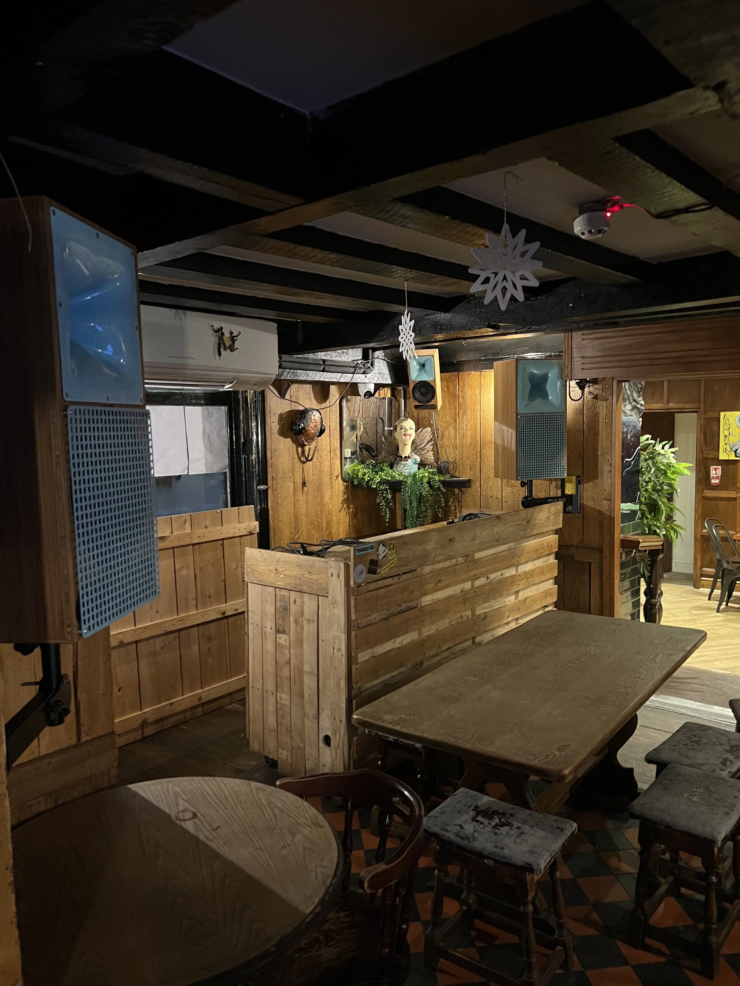 Interior of a cozy wooden restaurant or pub with tables, chairs, hanging snowflake decorations, and a small stage area with microphones and speakers.