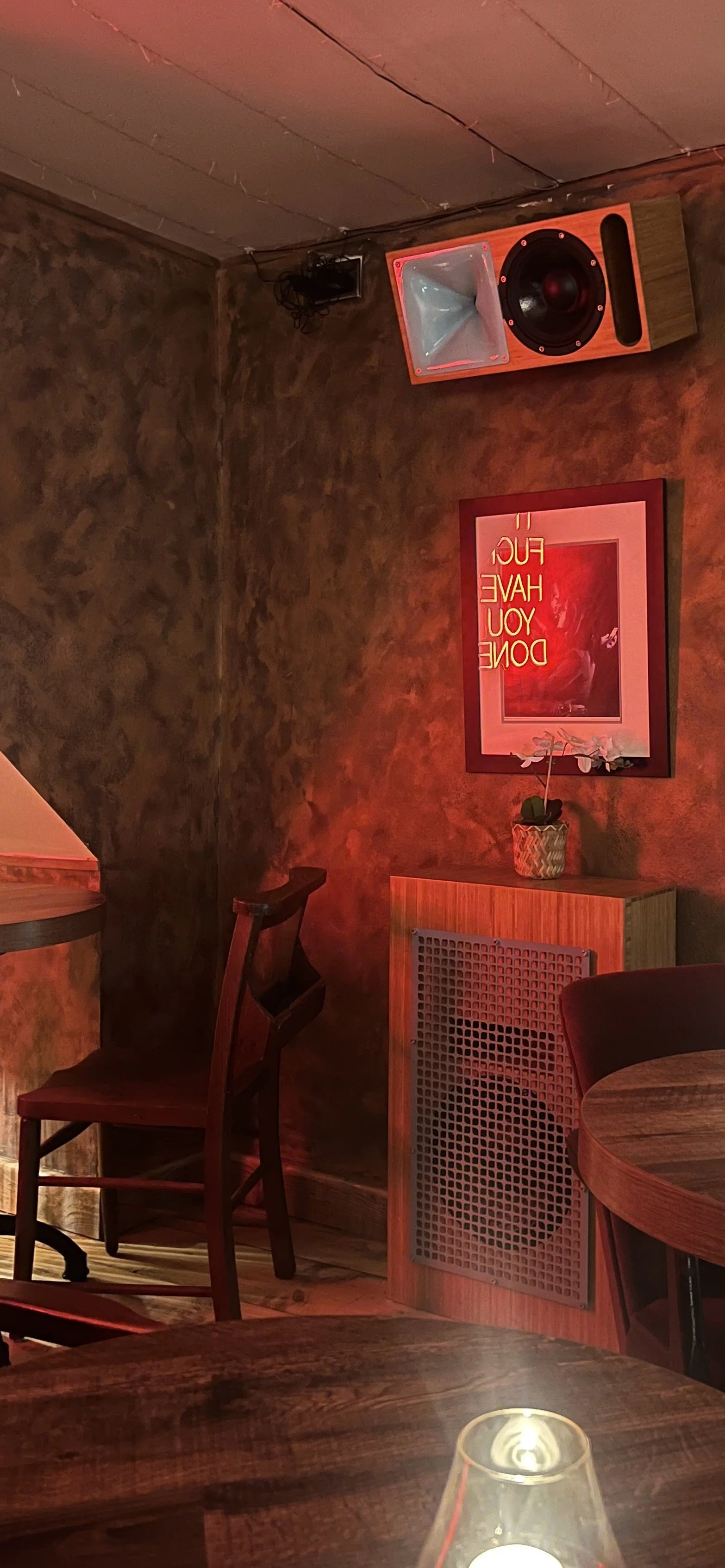A cozy restaurant or bar with textured dark walls, wooden furniture, and dim lighting. There is a framed sign on the wall that says, "IF YOU HAVE DON" with the last words cut off, and a small plant on a shelf below it. An illuminated speaker and sound equipment are mounted on the wall. A candle in a glass holder is on the table in foreground.