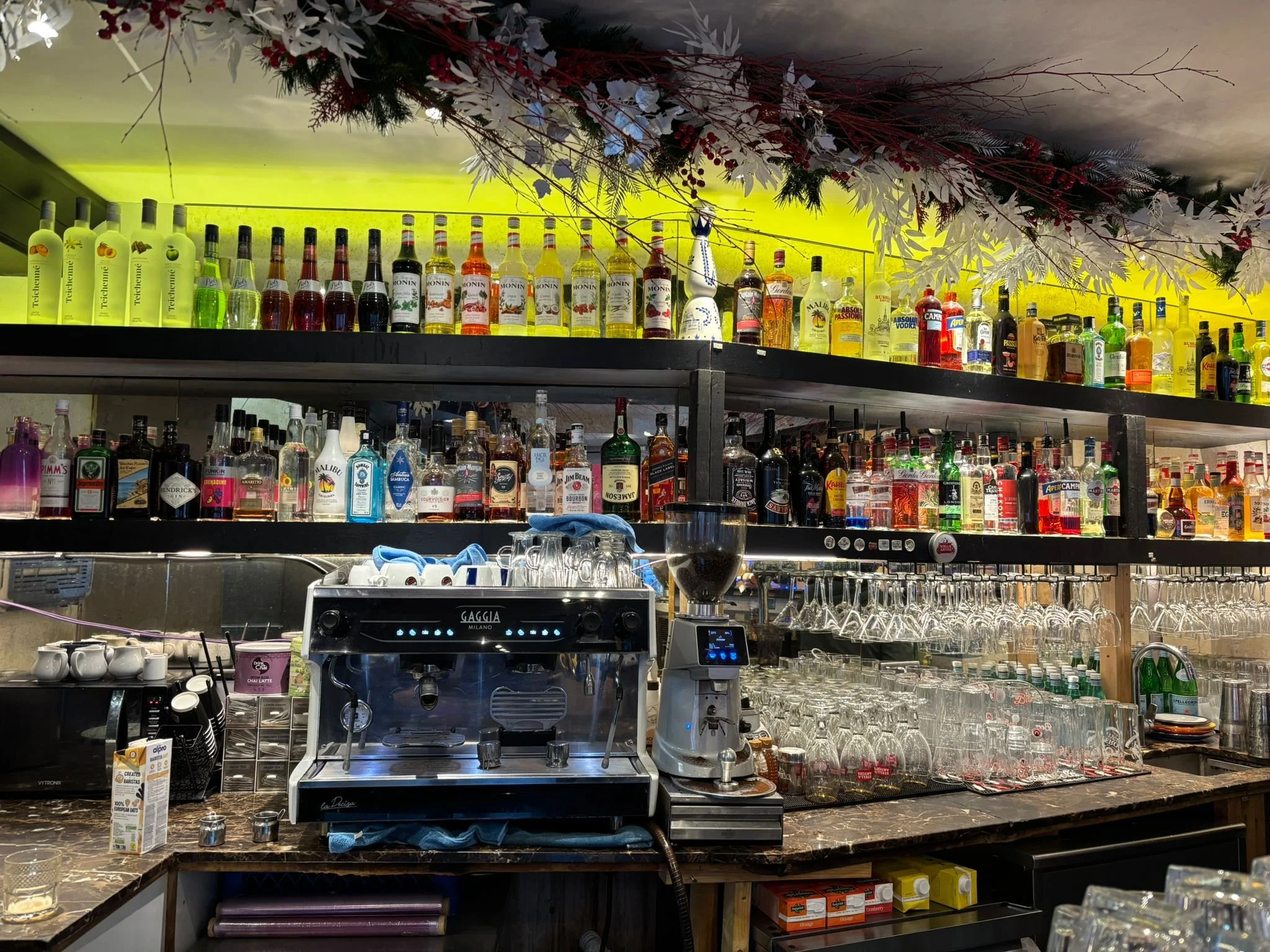 Bar with shelves stocked with various bottles of alcohol and glassware, with a coffee machine and blender on the countertop.