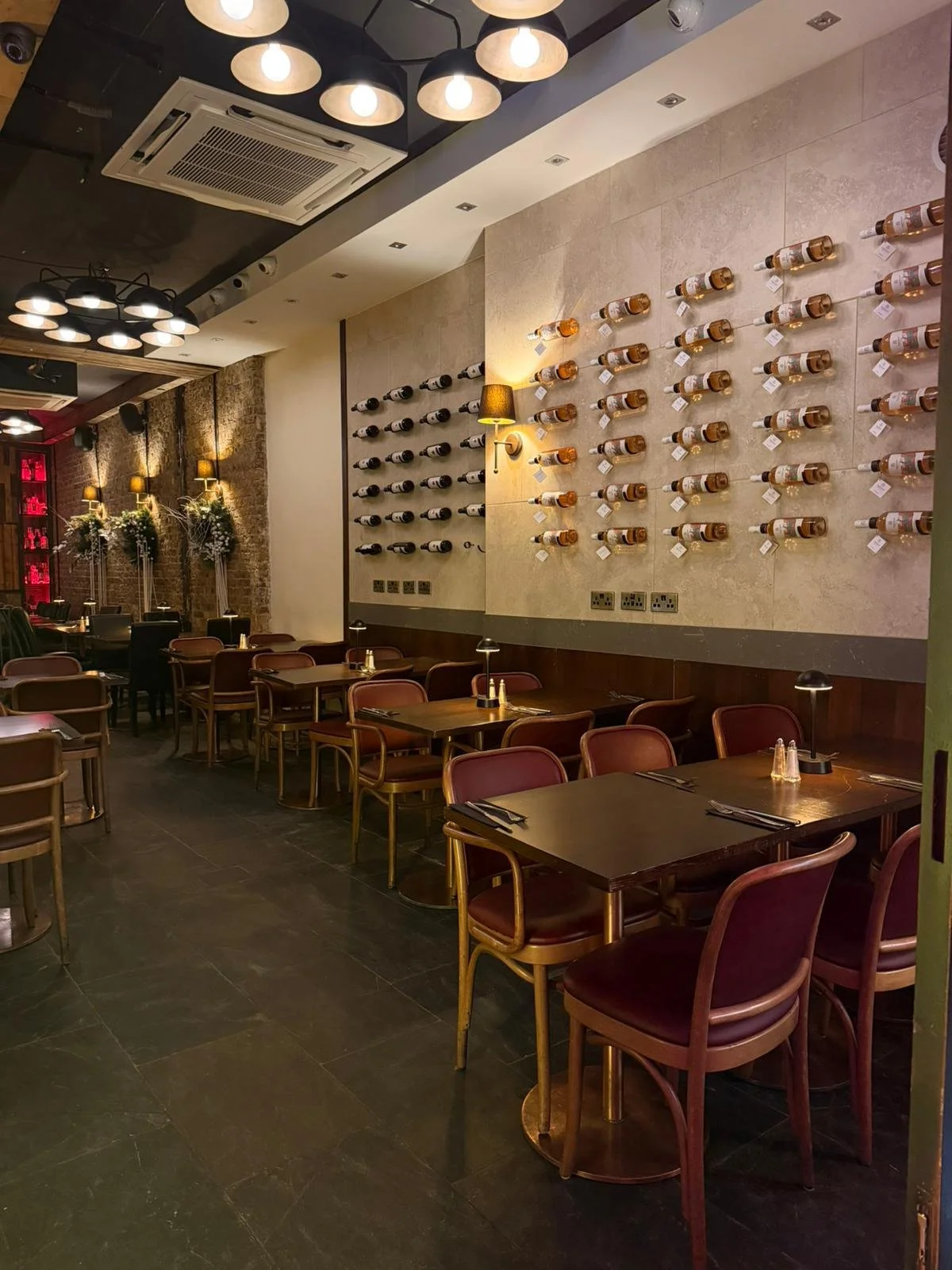 Interior of a restaurant with dark wood tables and chairs, wine bottles displayed on the wall, ambient lighting, and floral decorations in the background.