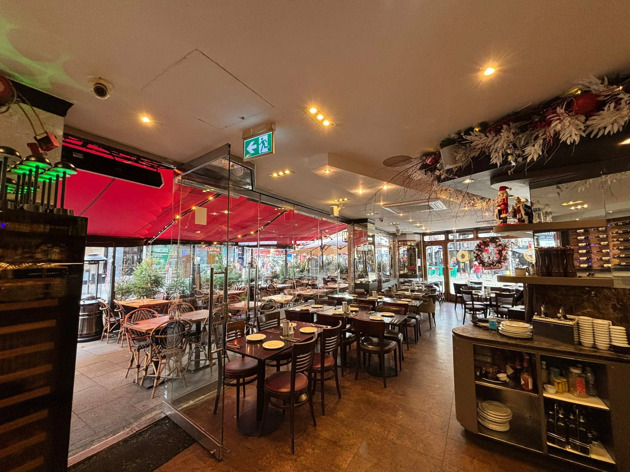 Empty restaurant with tables set for dining, decorated for Christmas, featuring festive wreaths, Christmas figures, and holiday garlands. Glass partition separates indoor and outdoor seating areas, with a street view outside.