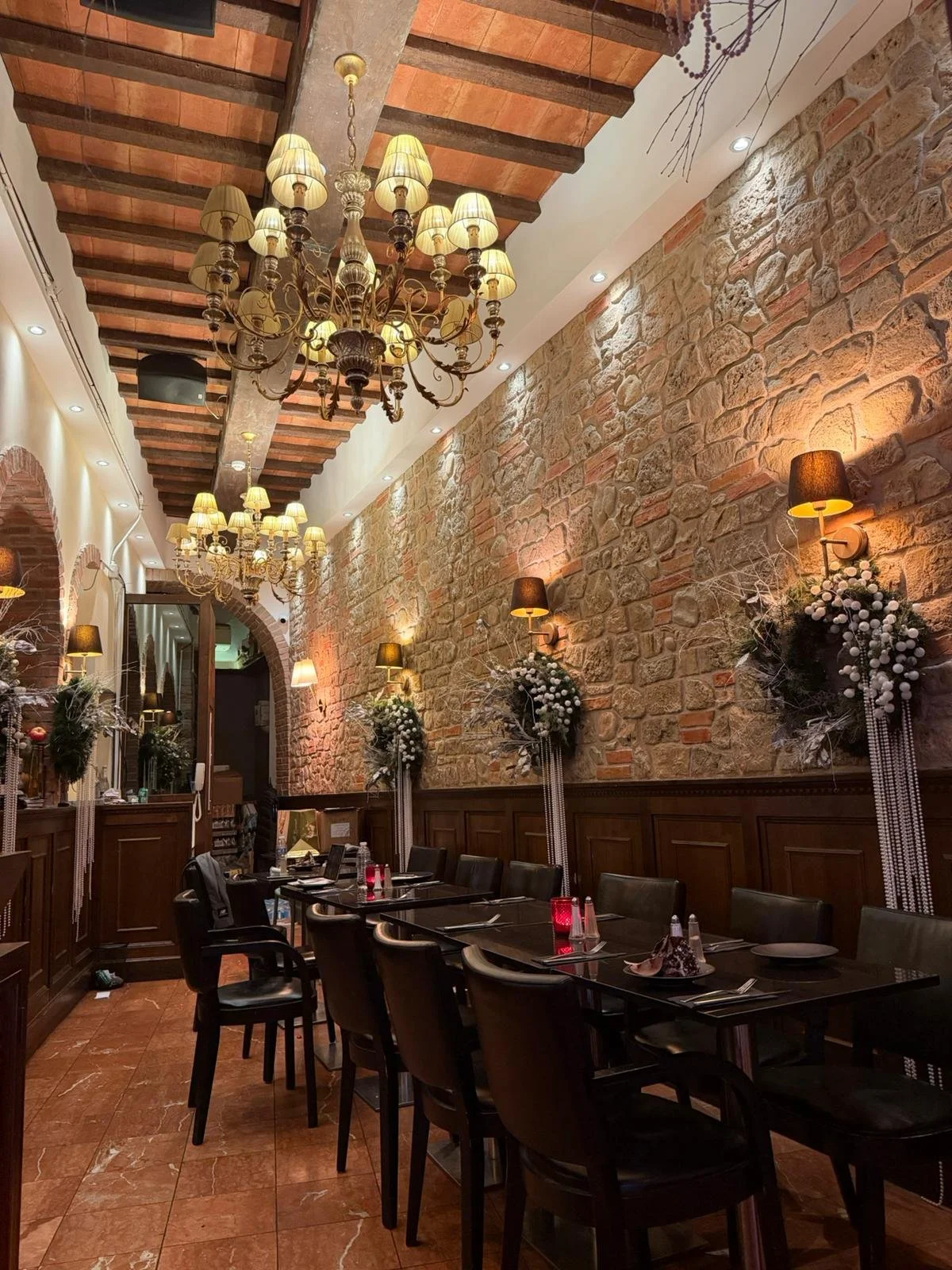 A cozy restaurant interior with a rustic stone wall, hanging chandeliers with multiple lights, wall-mounted black lamps, and floral decorations on the wall. A long dining table set with placemats, utensils, and wine glasses.