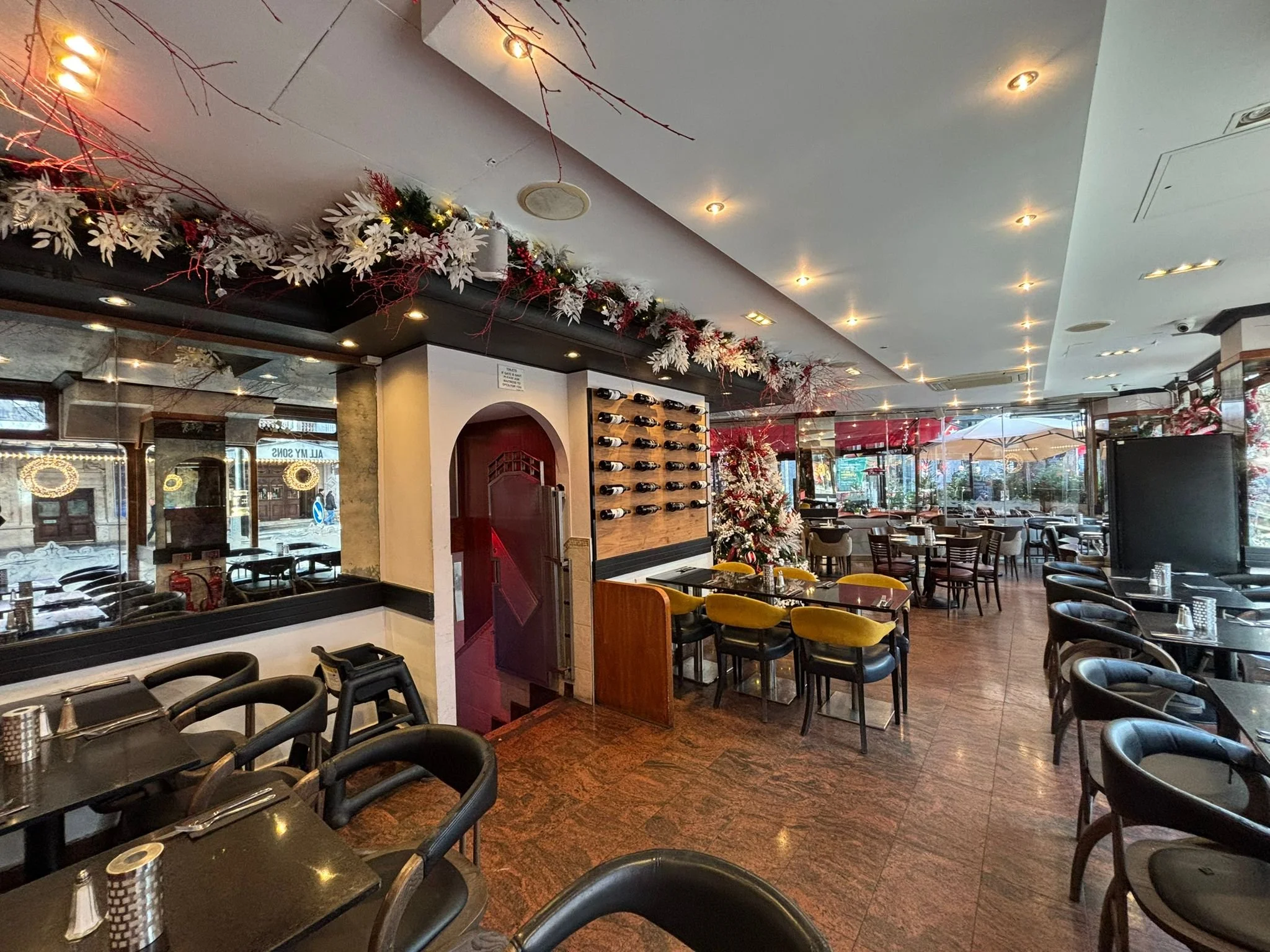 Empty restaurant with black tables, yellow and black chairs, decorated for Christmas with garlands, ornaments, and a Christmas tree, large windows, and indoor lighting.