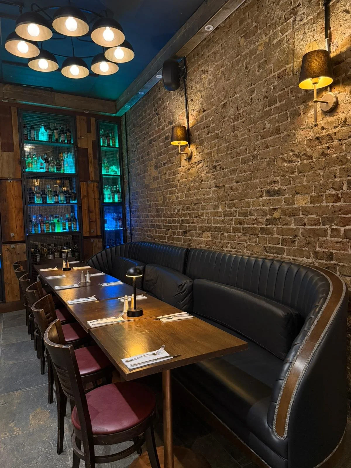 Interior of a restaurant with a long wooden table, red cushioned chairs, a black leather booth, exposed brick wall, and modern lighting fixtures.