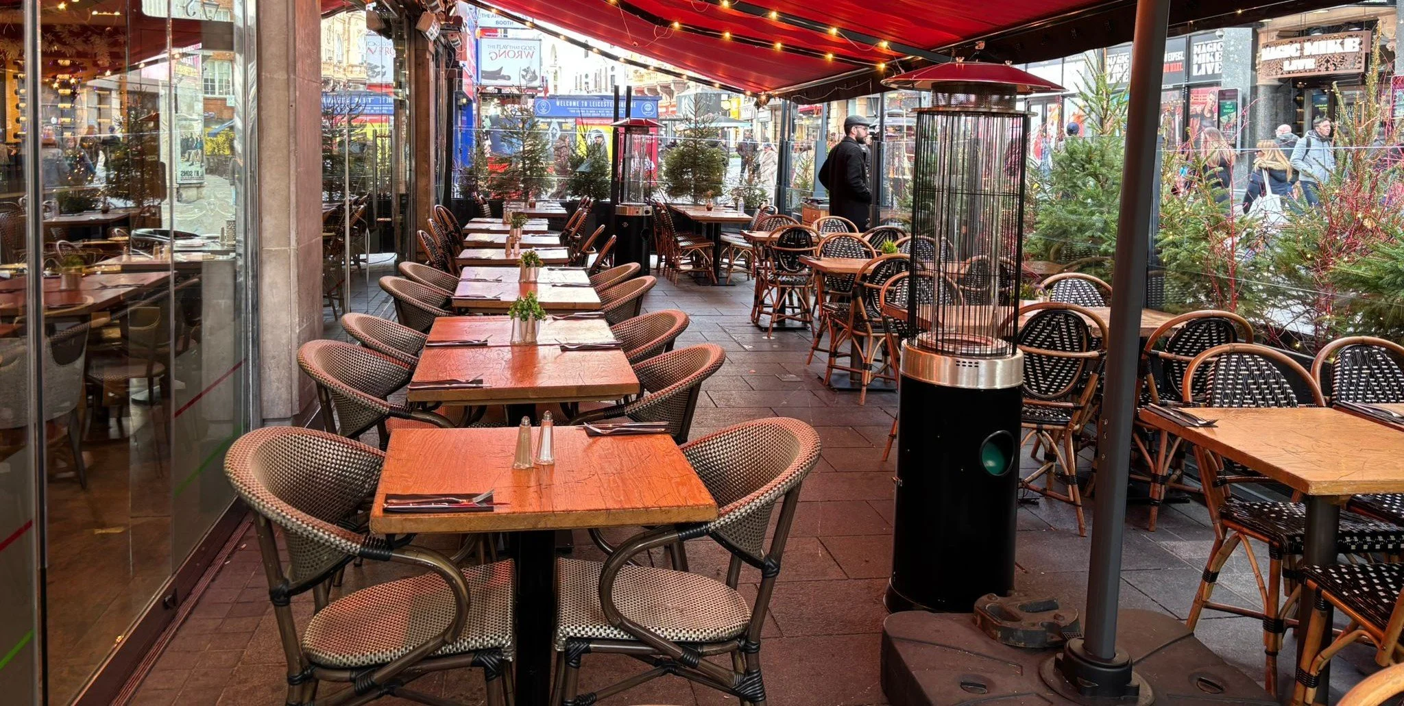Empty outdoor restaurant patio with wooden tables and woven chairs, heaters, and greenery, located on a bustling city street with pedestrians and storefronts visible in the background.