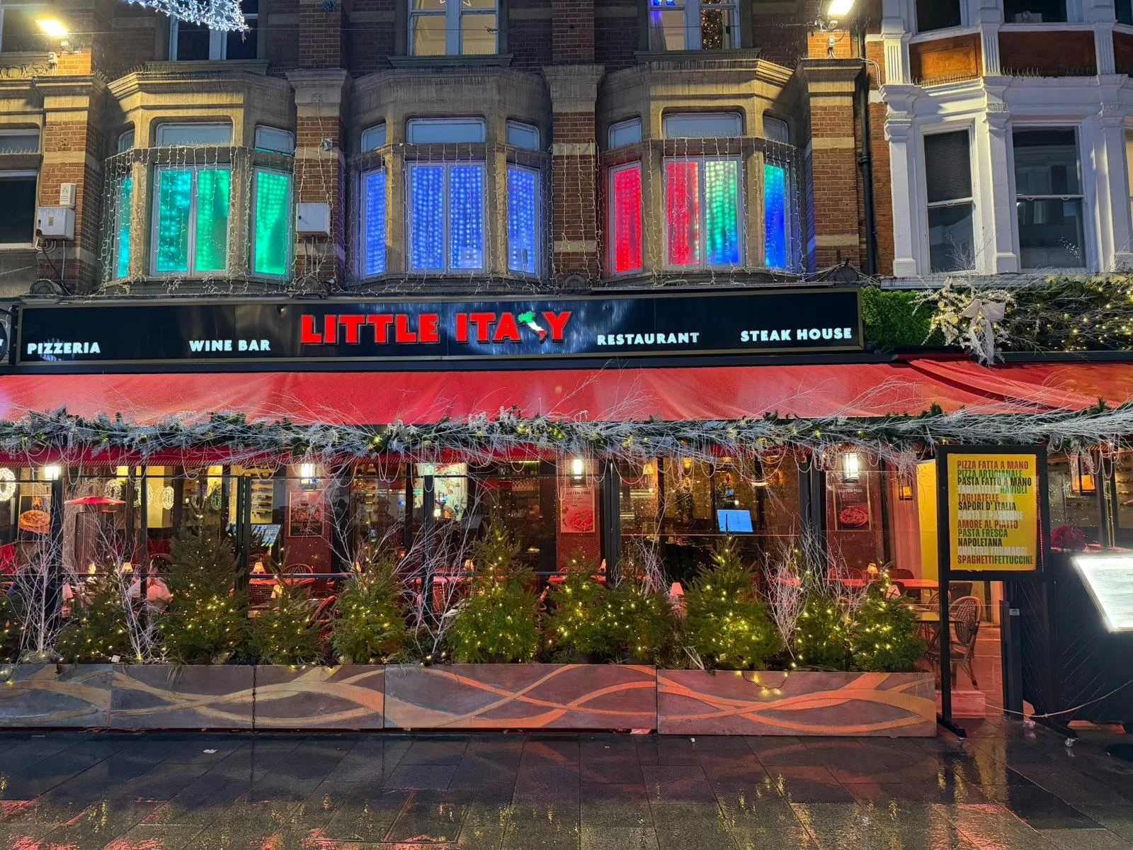 Exterior view of a restaurant named Little Italy decorated for Christmas with small Christmas trees, garlands, twinkling lights, and red awning, with colorful window lights on upper floor.