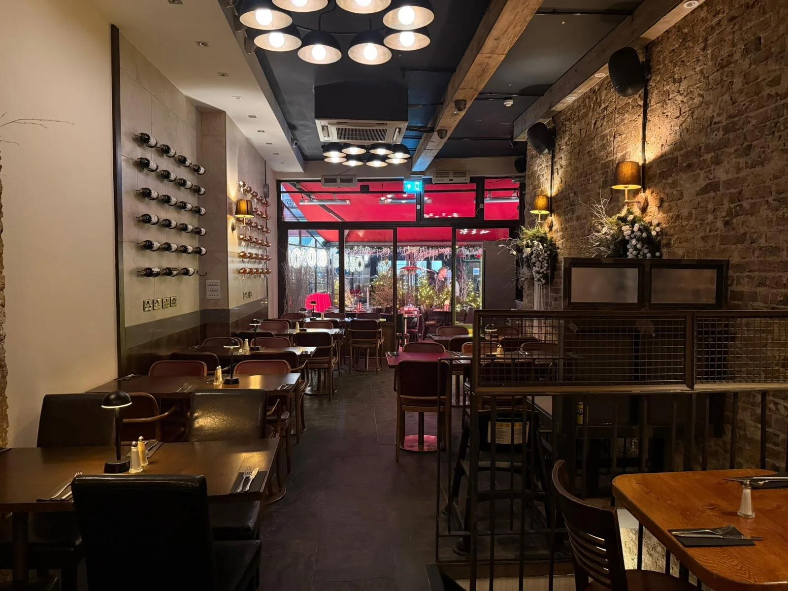 Interior of a cozy restaurant with brick and beige walls, tables set with utensils, and a view of an outdoor patio with red and white umbrellas.