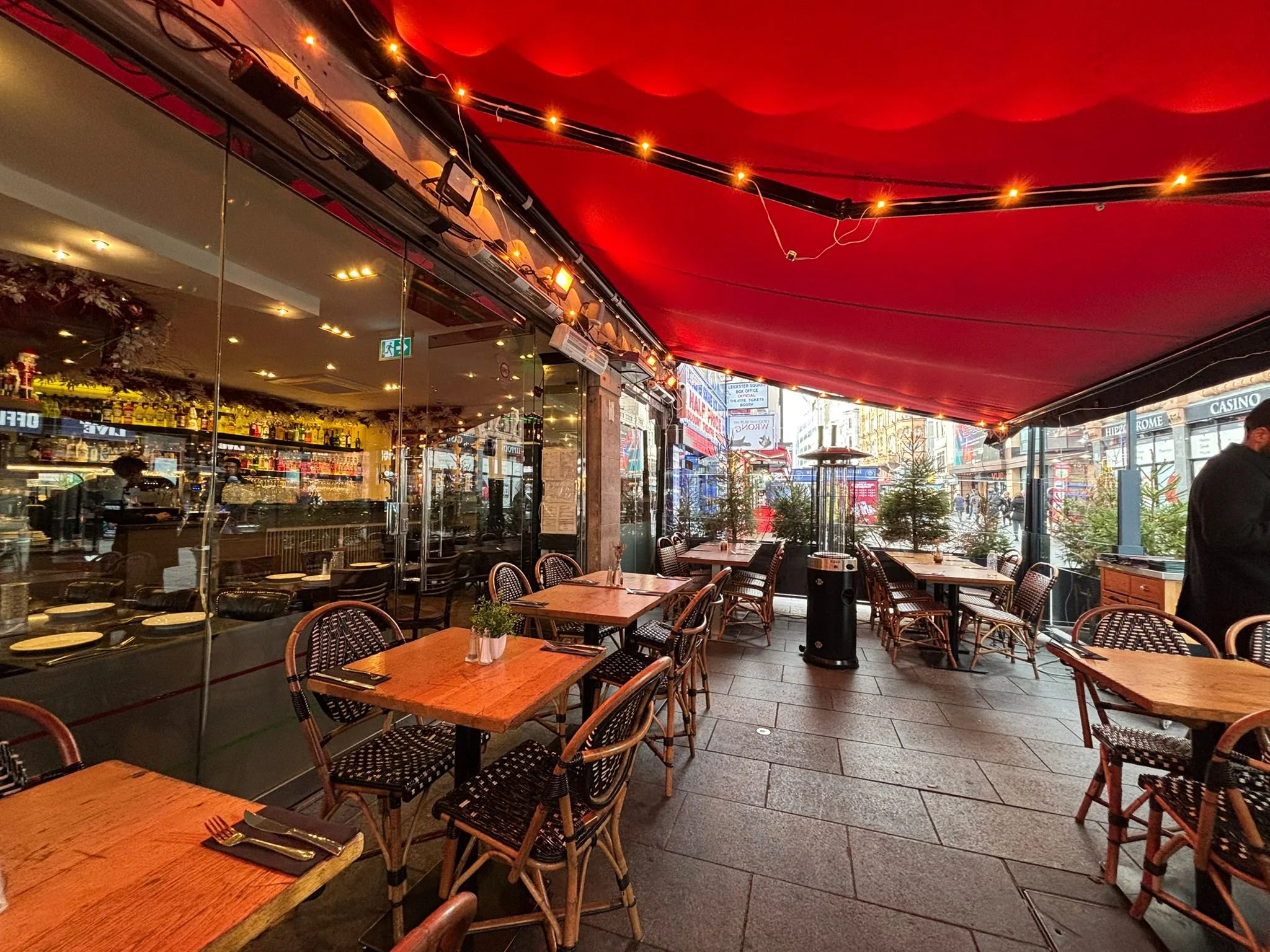 Empty outdoor patio of a restaurant with wooden tables, wicker chairs, string lights, and small potted plants, with a view of city street and storefronts.