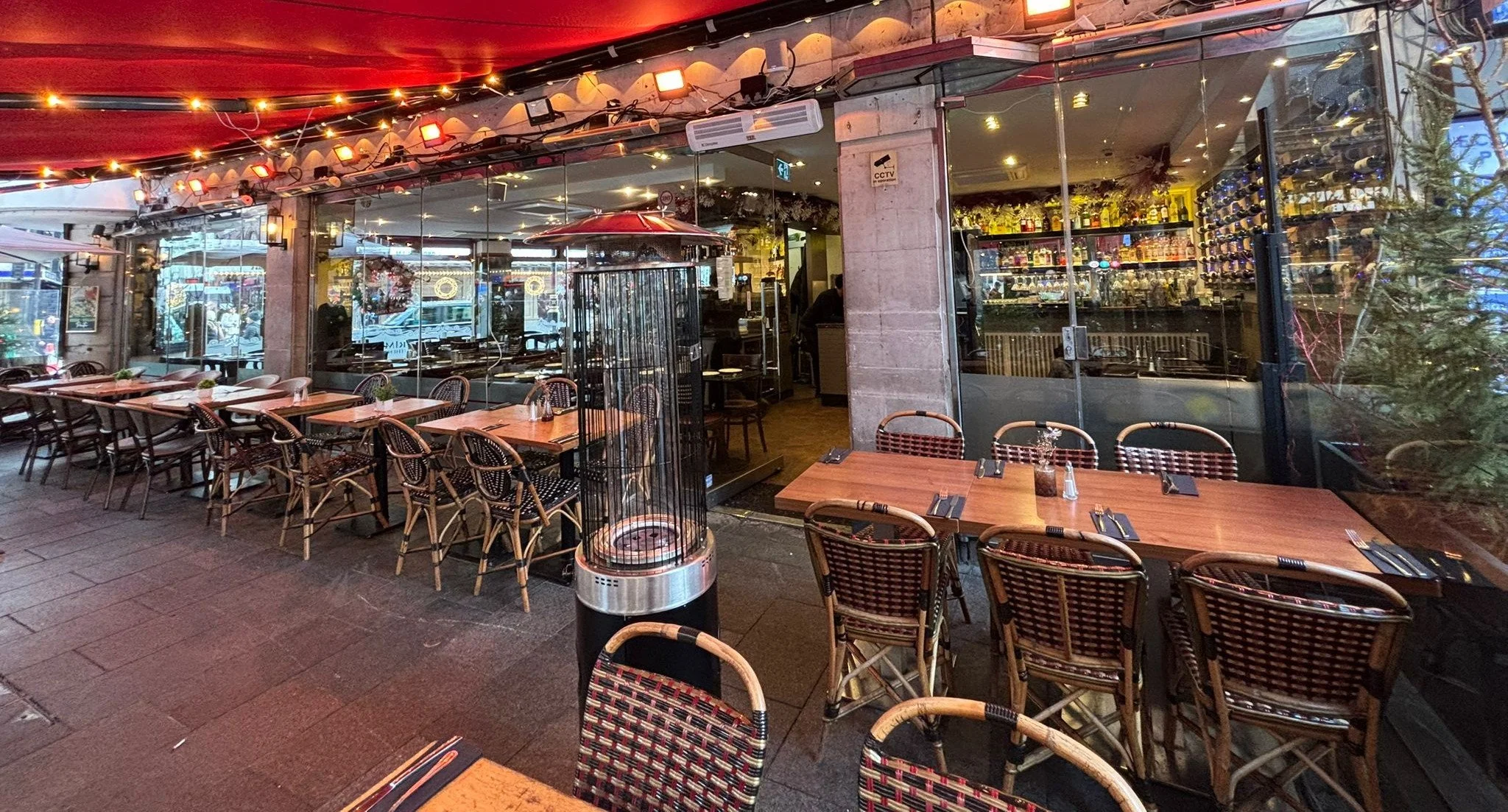 Outdoor restaurant patio with wooden tables, woven chairs, string lights, a patio heater, and a view of a bar inside through glass windows.
