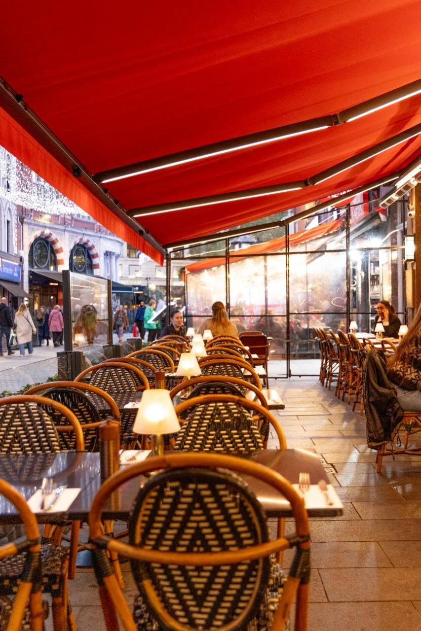 Outdoor restaurant seating area with woven chairs, tables set with cutlery, and small lamps, under a red awning; outside view of a busy street with pedestrians and storefronts.