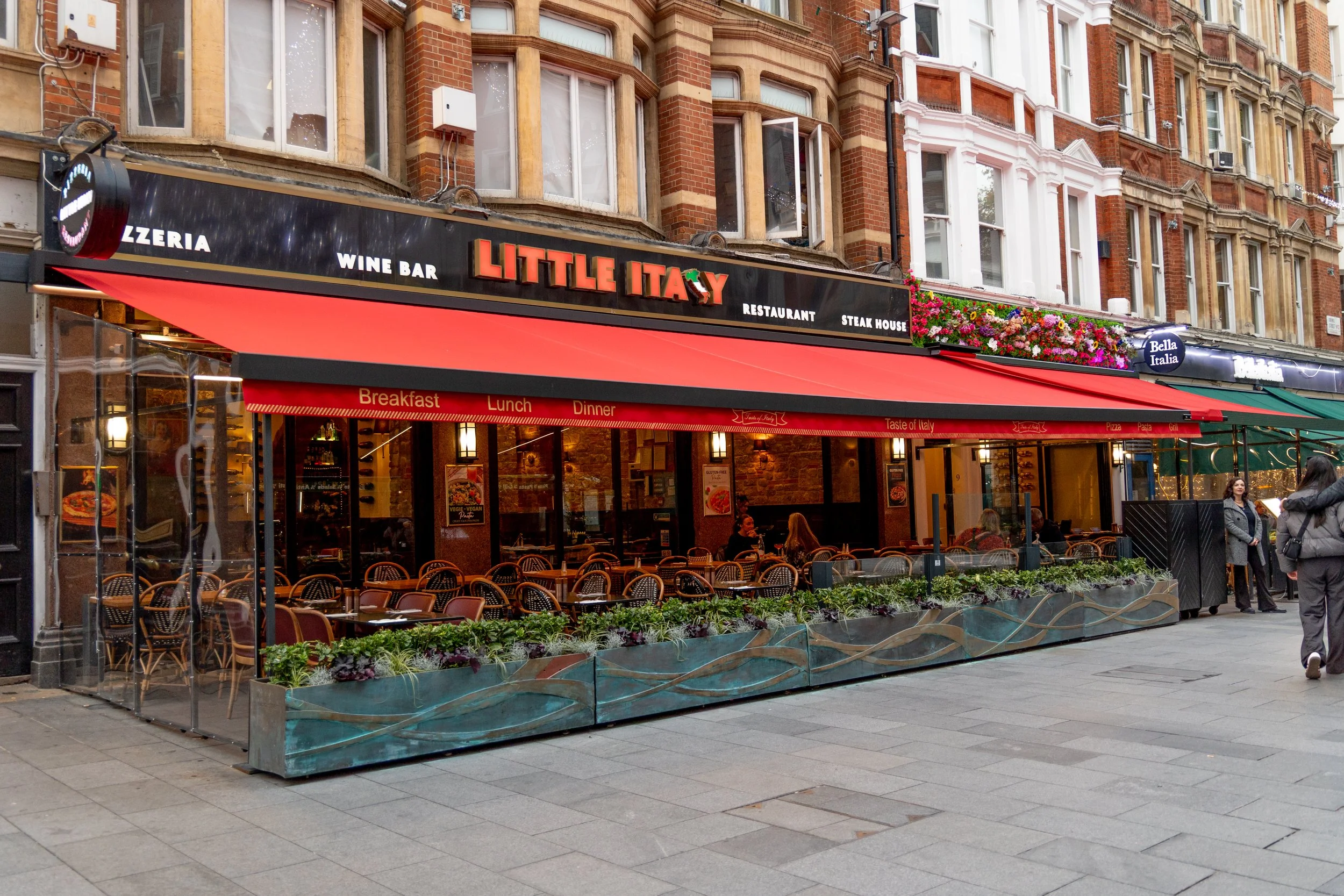Exterior view of a restaurant named 'Little Italy' with outdoor seating. The restaurant has a black sign with red and white lettering, a red awning, and outdoor tables and chairs. There are also advertisements for breakfast, lunch, and dinner on the 