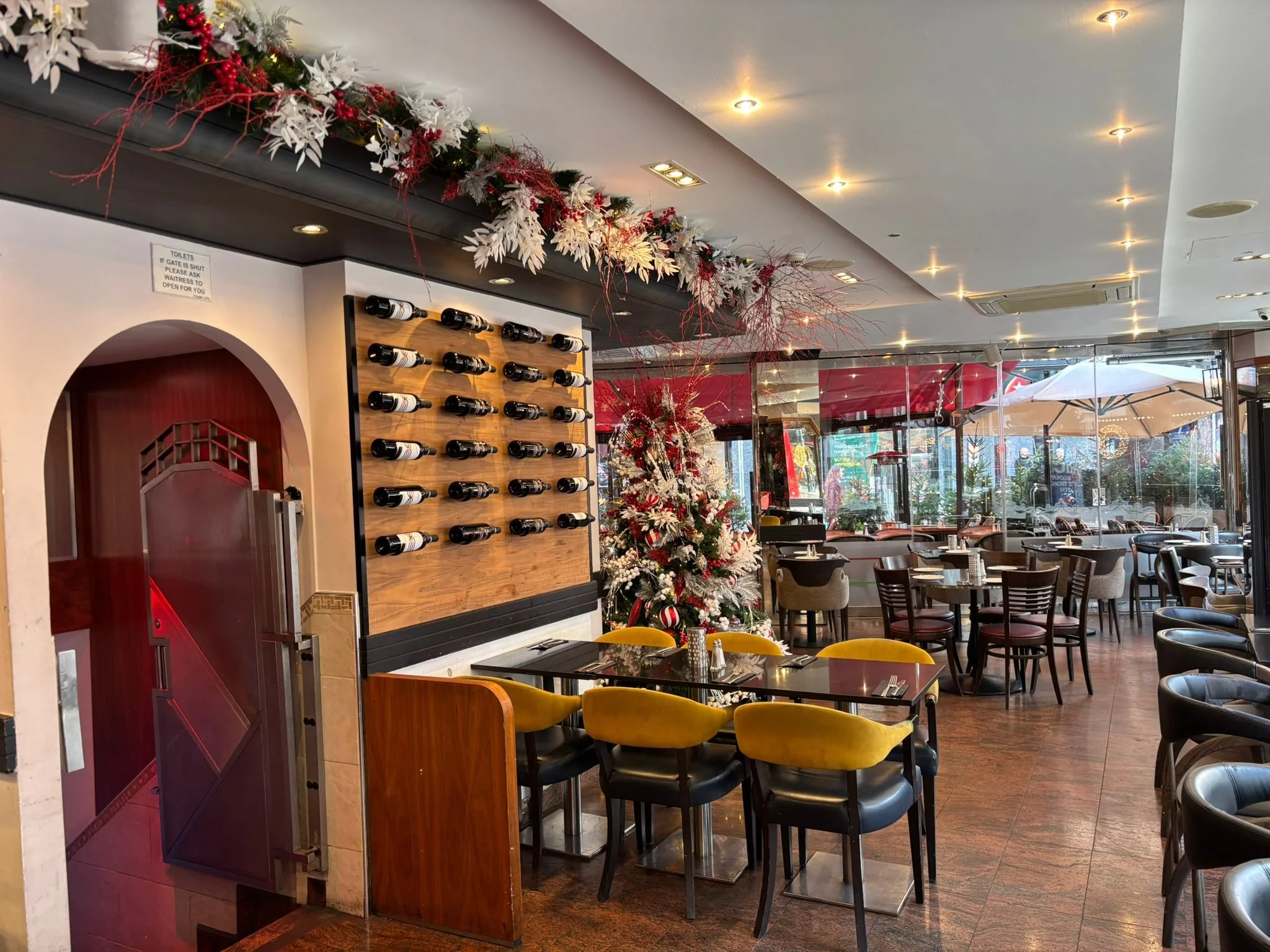 A restaurant decorated for Christmas with a white Christmas tree, a wine rack on the wall, and a garland of red and white ornaments hanging overhead. Patio seating with umbrellas is visible outside.