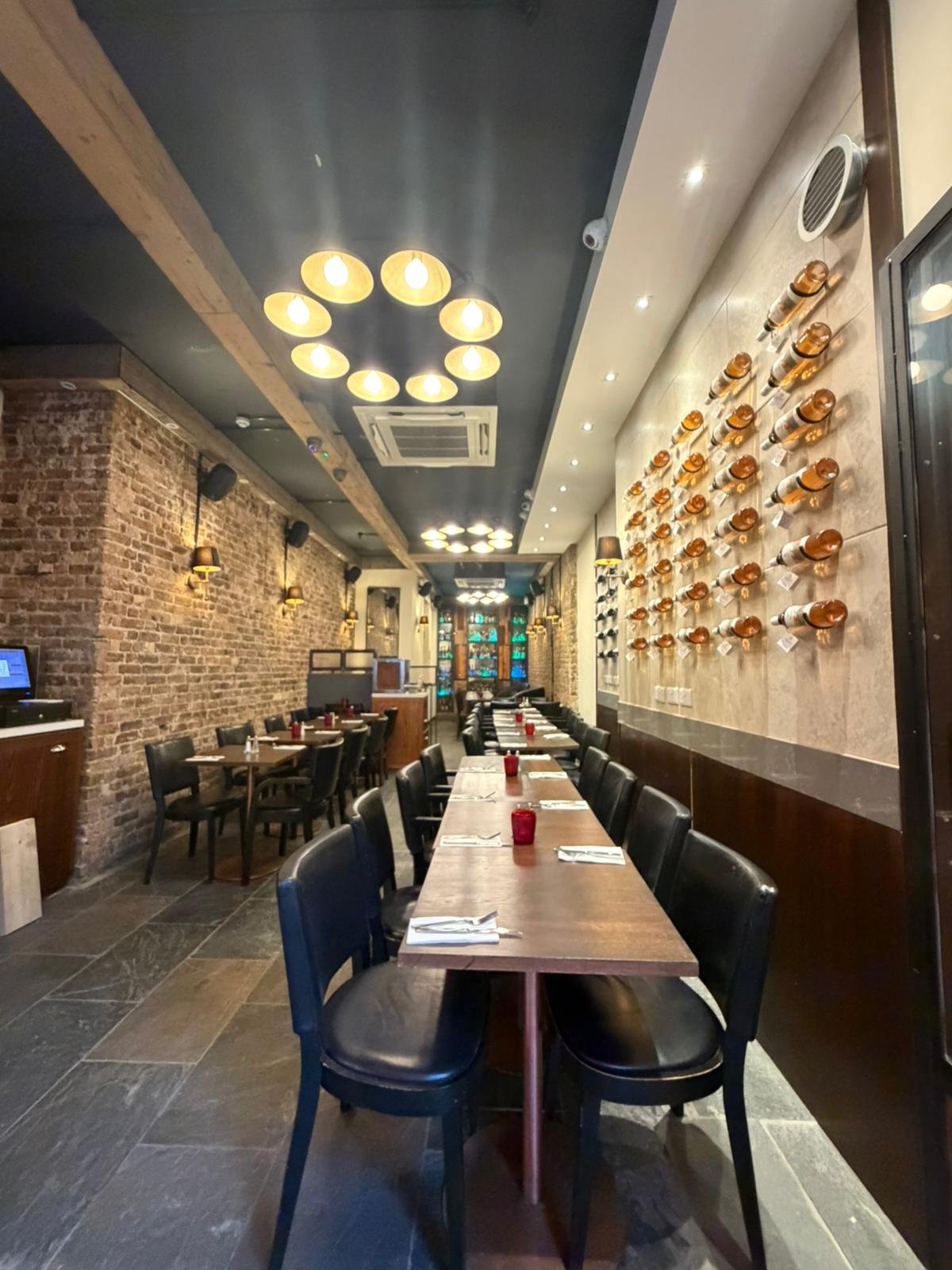 Interior of a modern restaurant with brick walls, wooden beams, and a wine display on the wall. Wooden tables are set with napkins and red candle holders, and black chairs are arranged around the tables.