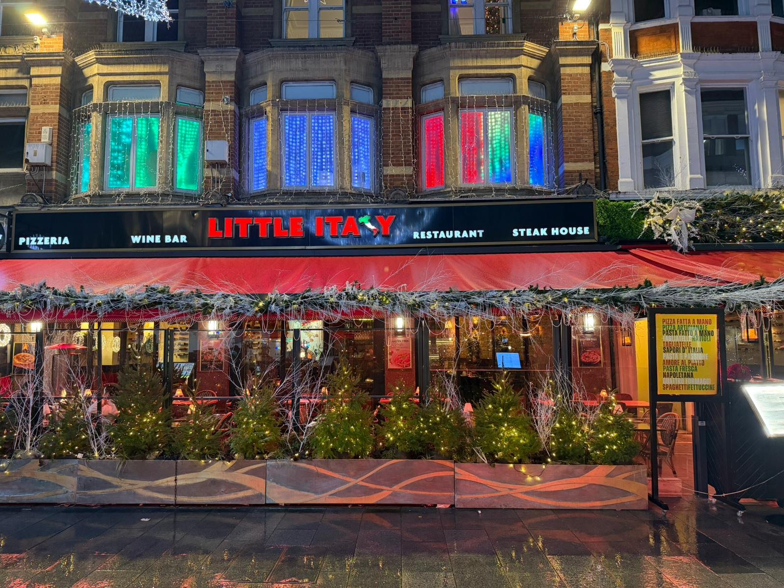 Exterior of an Italian restaurant named Little Italy decorated with Christmas lights, wreaths, small Christmas trees, and colorful neon window displays.