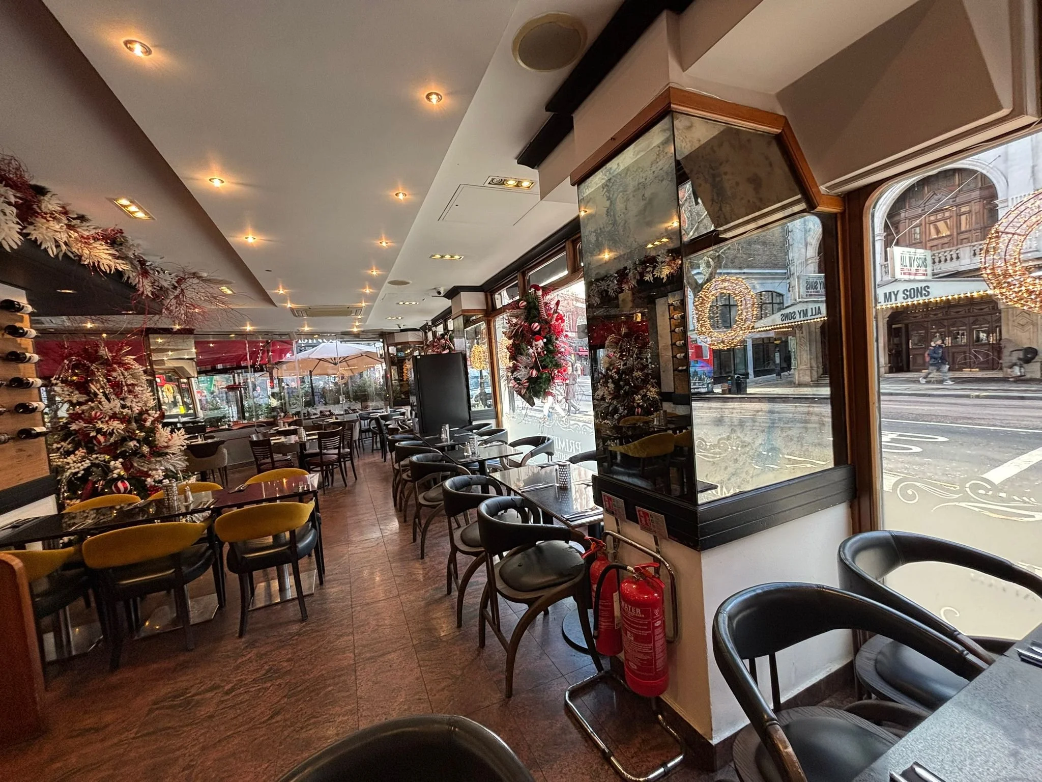 Interior of a restaurant decorated for Christmas with Christmas trees, wreaths, and holiday ornaments, featuring tables and chairs along large windows with street view.