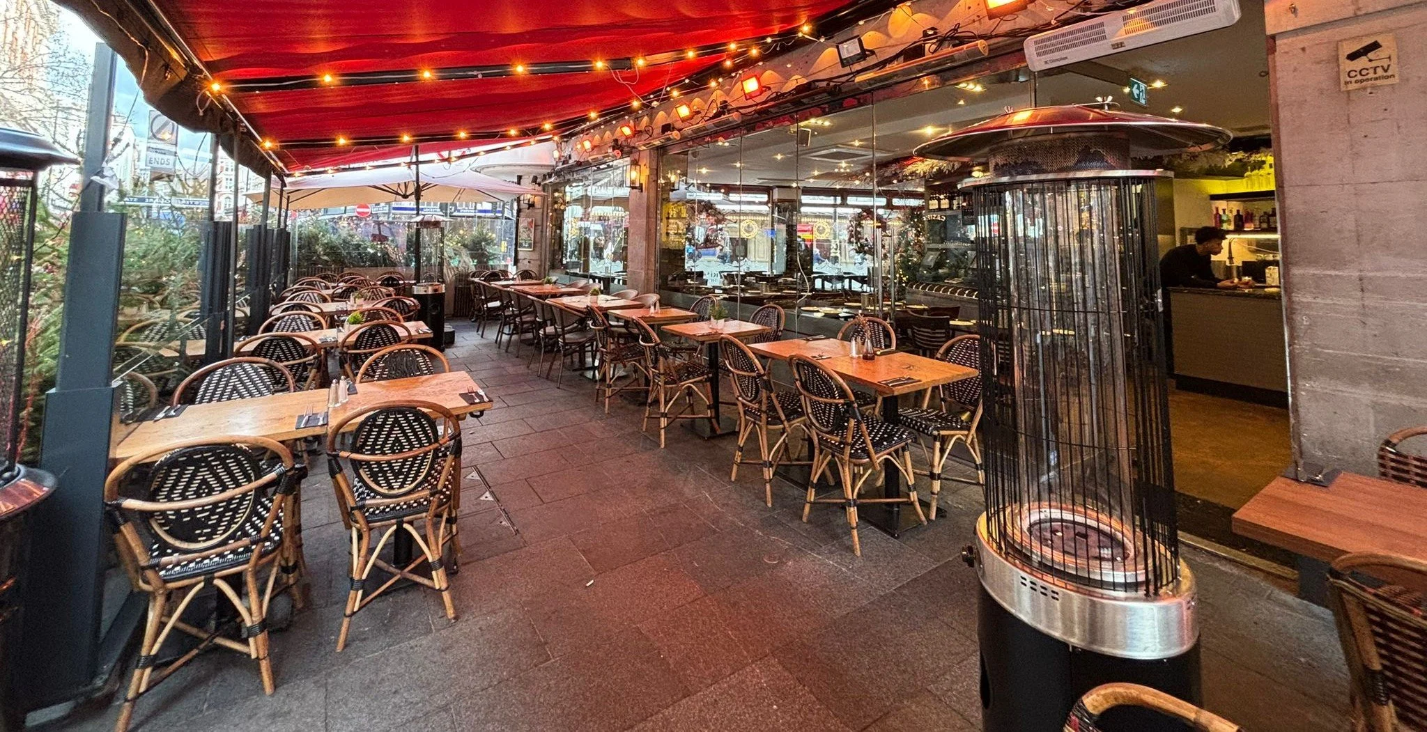 Empty outdoor dining area with wooden tables, rattan chairs, string lights, and a tall patio heater, with a view of street outside.