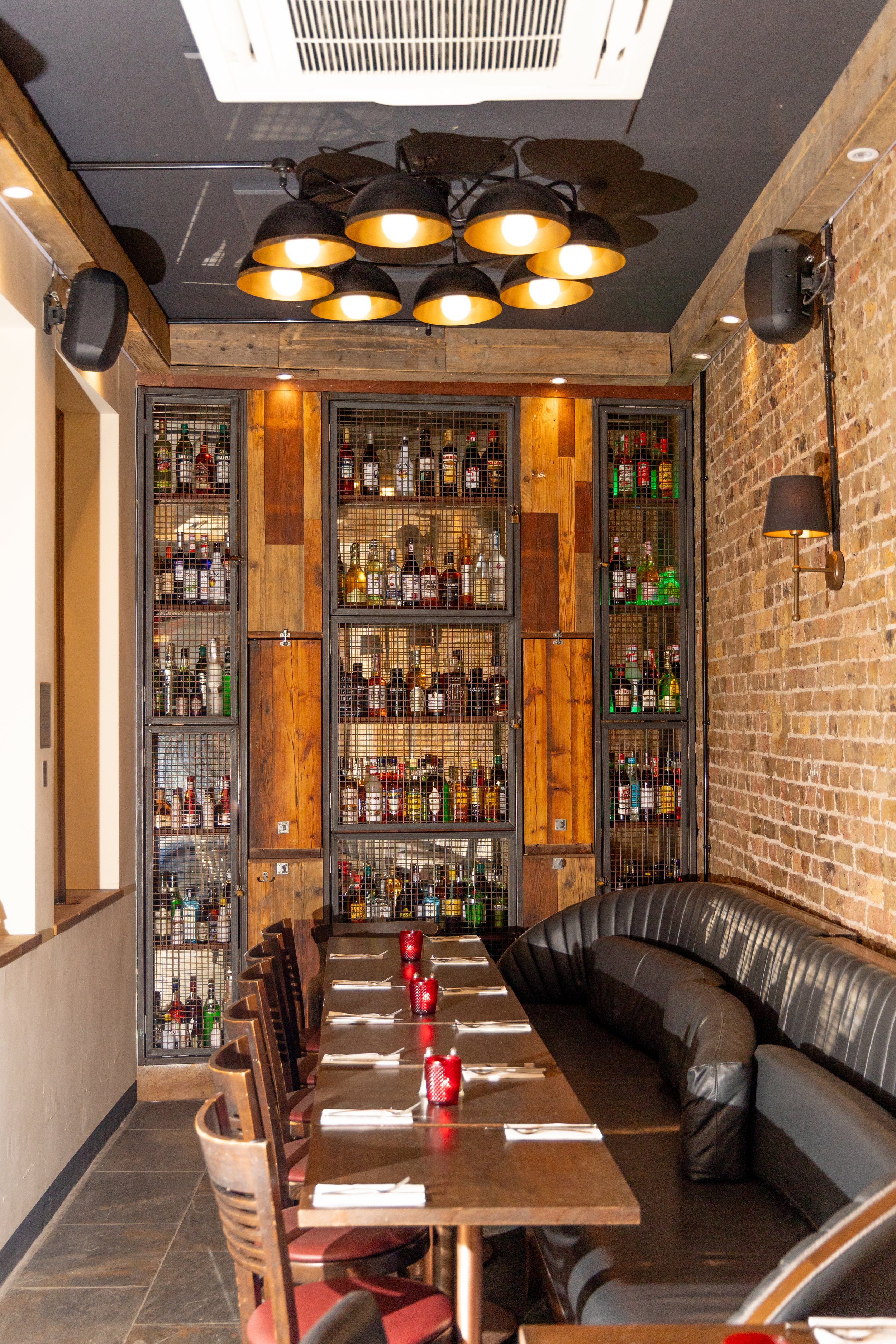 A dining area with a wooden table set with red candle holders, surrounded by wooden chairs and a black leather banquette. A large display of liquor bottles in metal cages is on the back wall, and a modern chandelier hangs from the ceiling overhead. T