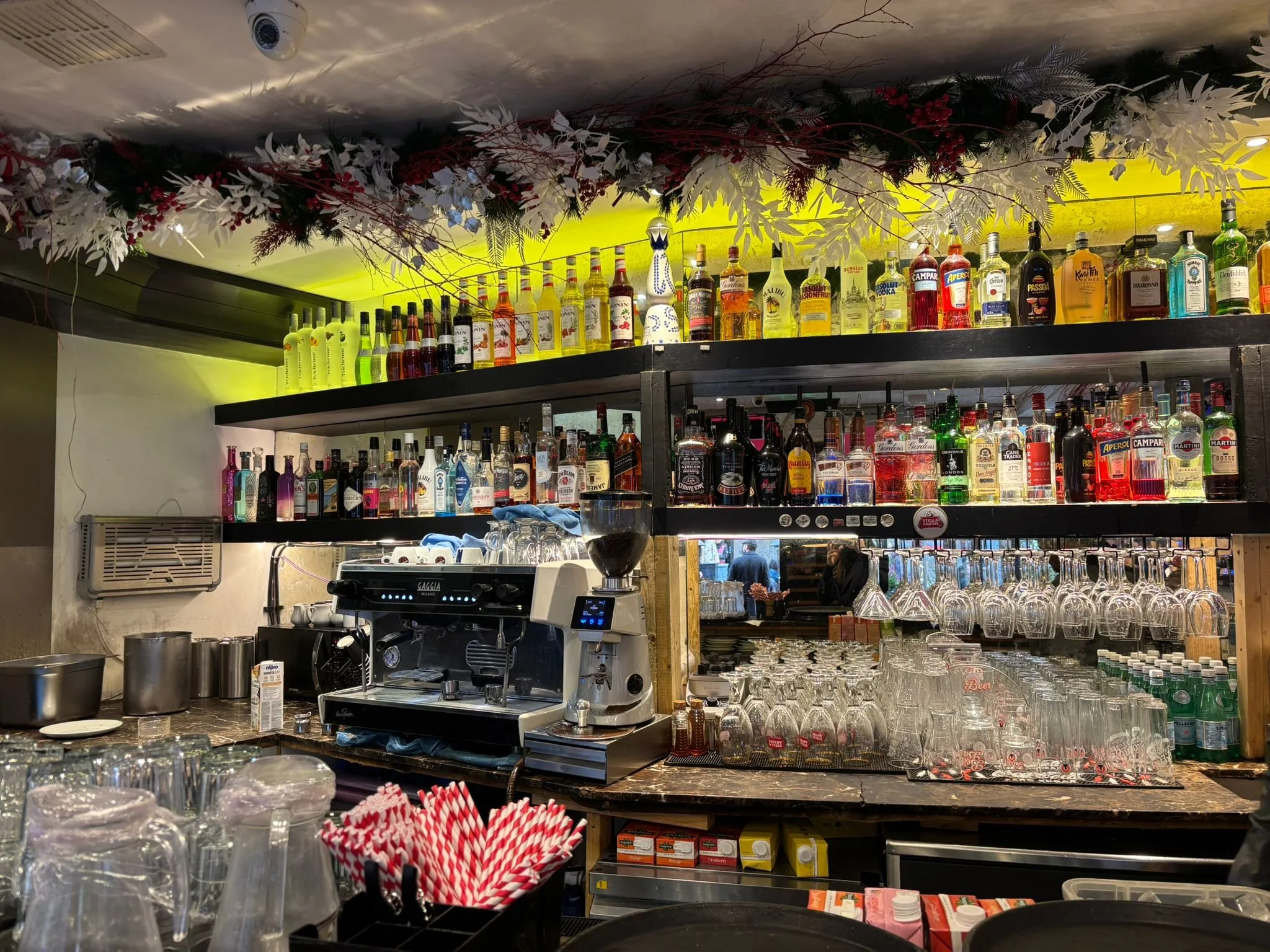 Bar area with shelves of alcohol bottles, glasses, and coffee machine, decorated with holiday garlands and flowers.