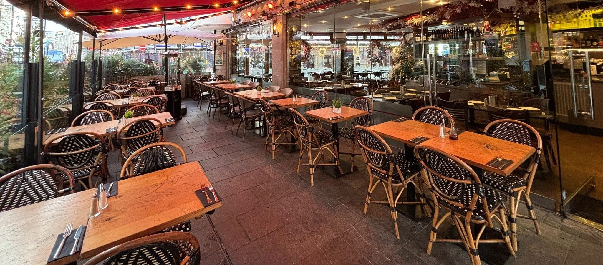 Empty outdoor patio of a restaurant with wooden tables, black wicker chairs, some with black and white cushions. Tables are set with utensils, salt and pepper shakers, and small potted plants. Large glass windows separate the patio from the indoor di