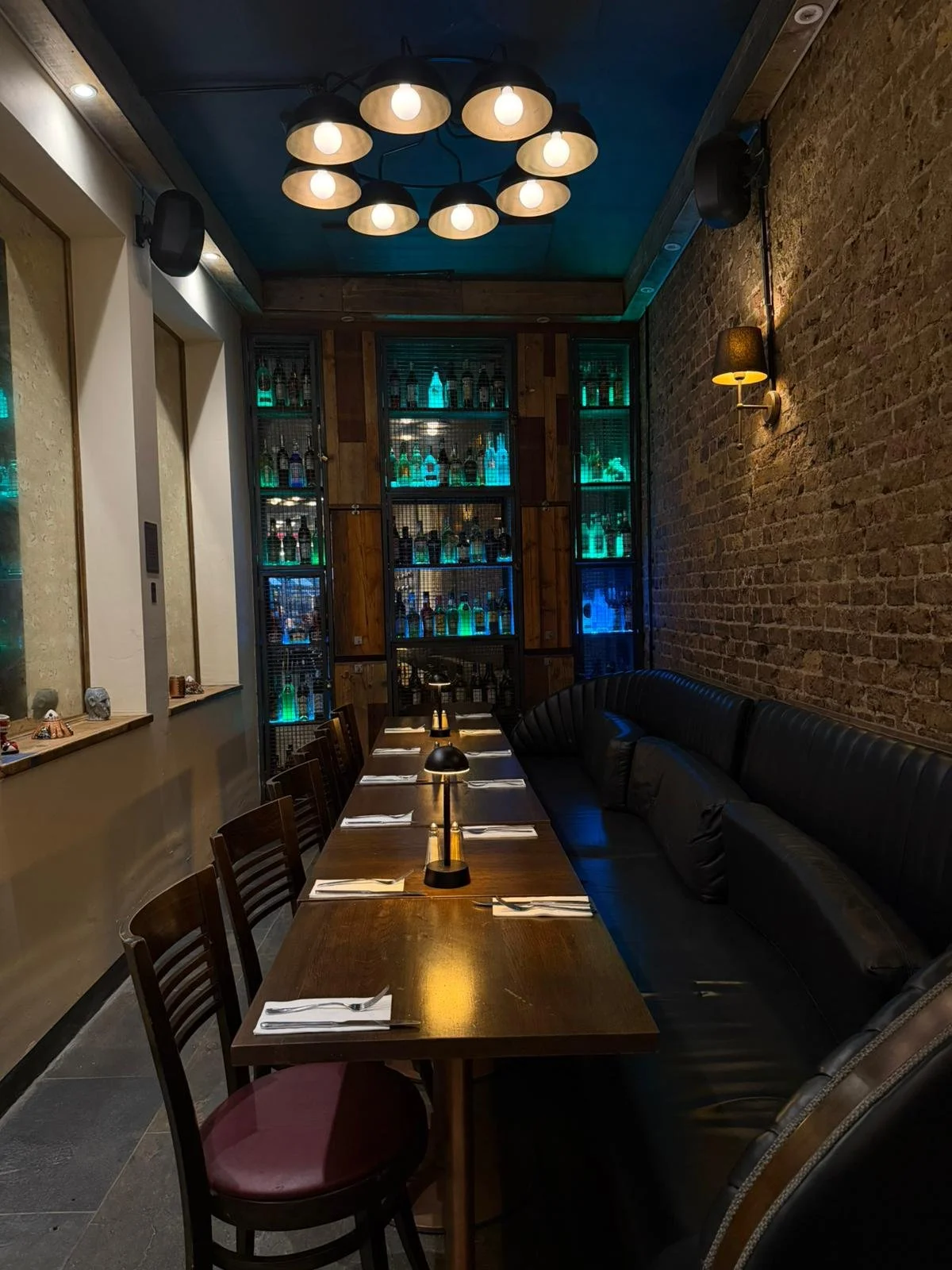 Dimly lit restaurant interior with a long wooden table set for six, black chairs, and a curved black leather bench on the right. A modern black chandelier with exposed bulbs hangs from the ceiling. A brick wall on the right side, and a wooden shelvin