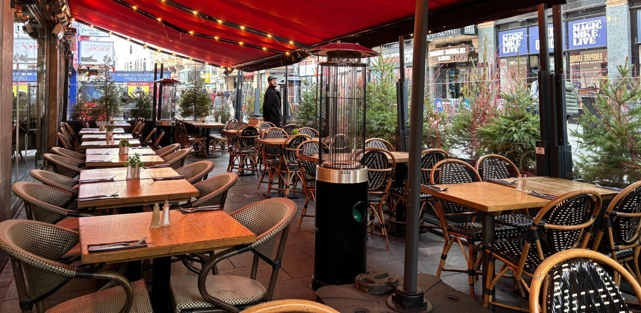 Indoor restaurant seating area with empty tables and chairs, view of outside street scene through large windows, decorated with Christmas trees and festive lights.