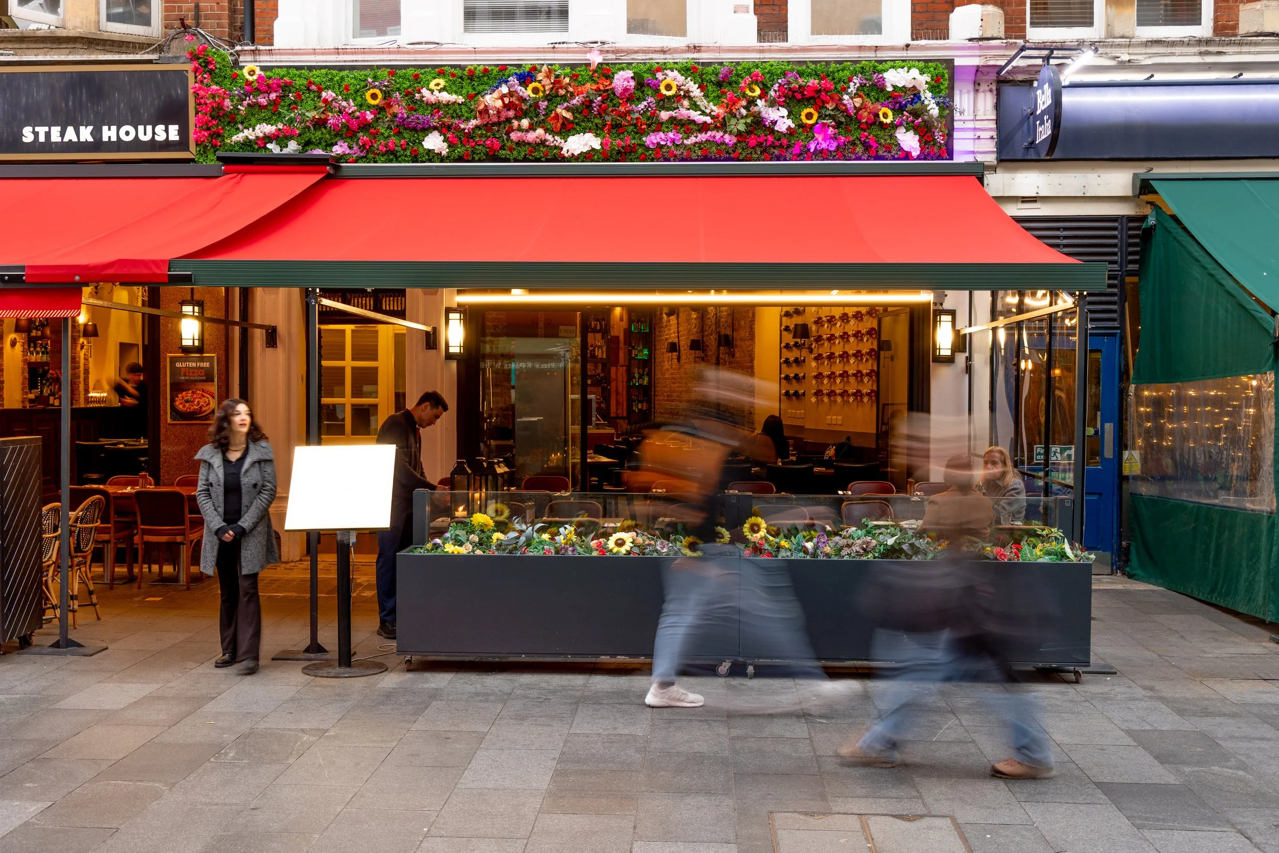 Street view of a restaurant with a red awning, large flower arrangement above, and a window revealing the interior where people are dining and staff are working. Pedestrians are walking by, some blurred due to motion.