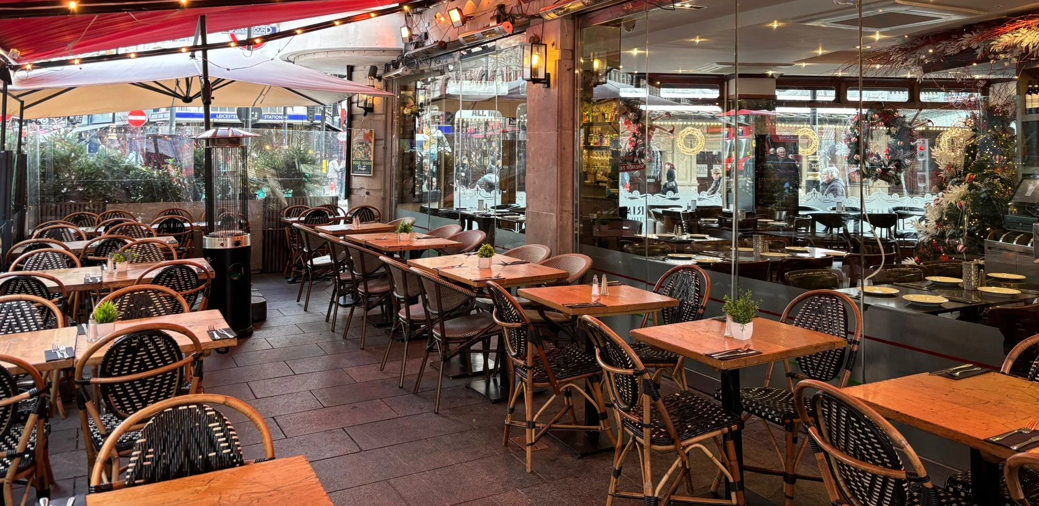 Outdoor restaurant seating area with wooden tables, black and brown chairs, small potted plants, and a heater, with a glass wall decorated for Christmas and a view of the city street outside.
