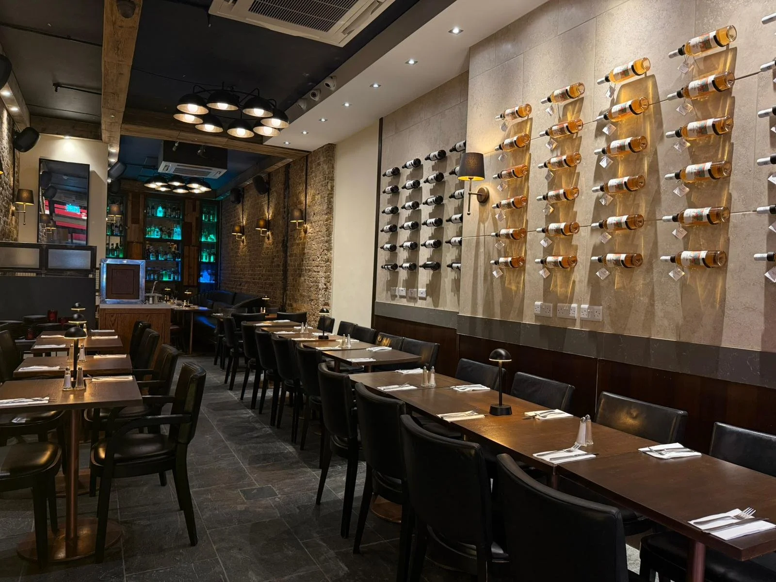 Interior of a modern restaurant with dark wooden tables, black chairs, a wine display on the wall, and ambient lighting.