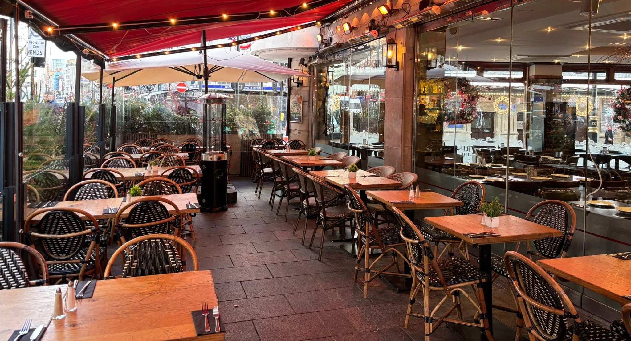 Empty outdoor patio of a restaurant with wooden tables, black and woven chairs, potted plants, and string lights, enclosed by glass panels with a city street visible outside.