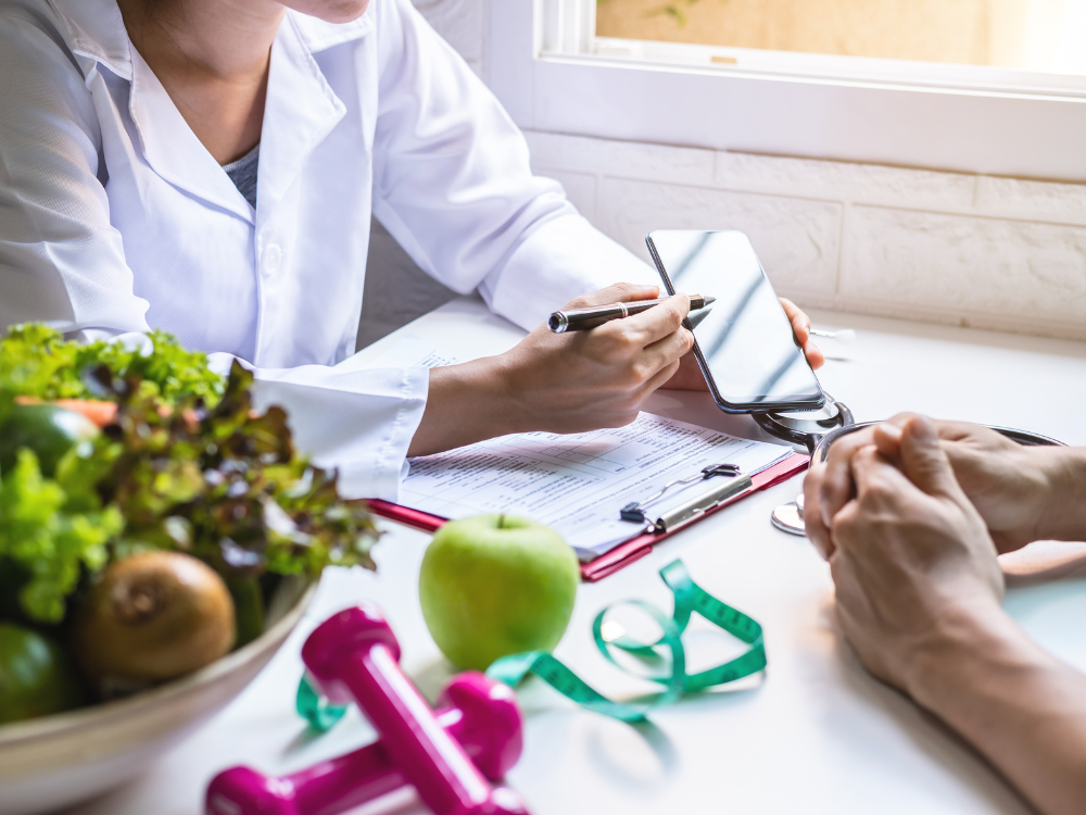 Eine Ärztin führt ein Beratungsgespräch mit einer Patientin in einer Klinik oder Praxis. Auf dem Tisch liegen frisches Obst, ein Blutdruckmesser, ein rosa Wasserflasche, eine Akte und ein Tablet.