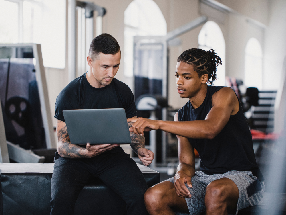 Zwei Männer im Fitnessstudio, die auf einen Laptop schauen, einer zeigt auf den Bildschirm, beide tragen sportliche Kleidung.