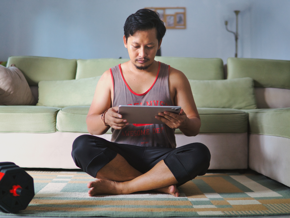 Mann sitzt im Wohnzimmer, liest auf einem Tablet, umgeben von einer grünen Couch, Kissen und einem Teppich.