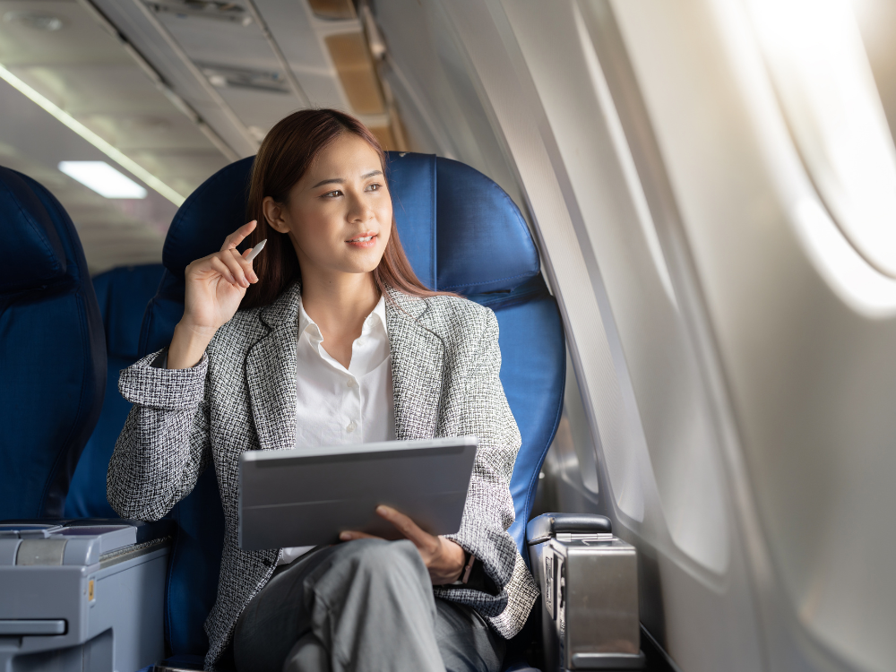 Frau sitzt in einem Flugzeug und schaut aus dem Fenster, während sie ein Tablet hält.