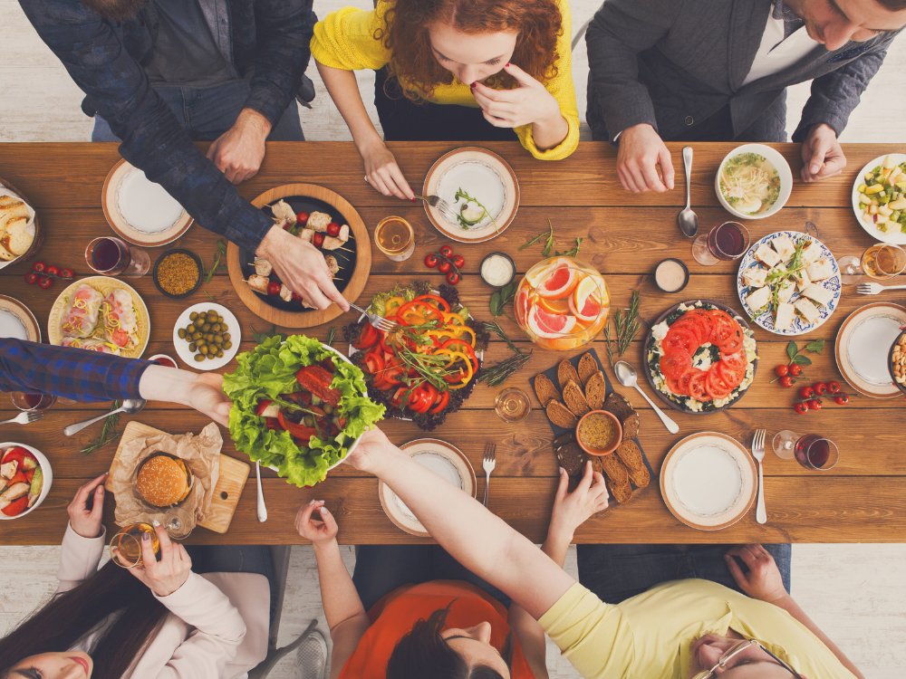 Blick von oben auf eine festliche Mahlzeit mit verschiedenen Speisen und Getränken auf einem Holztisch, umgeben von mehreren Personen, die gemeinsam essen und sprechen.