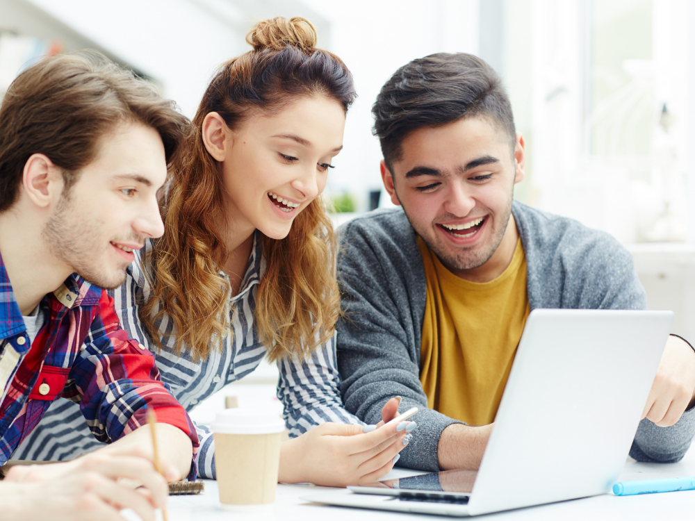 Drei junge Menschen schauen lachend auf einen Laptop in einem hellen Raum.
