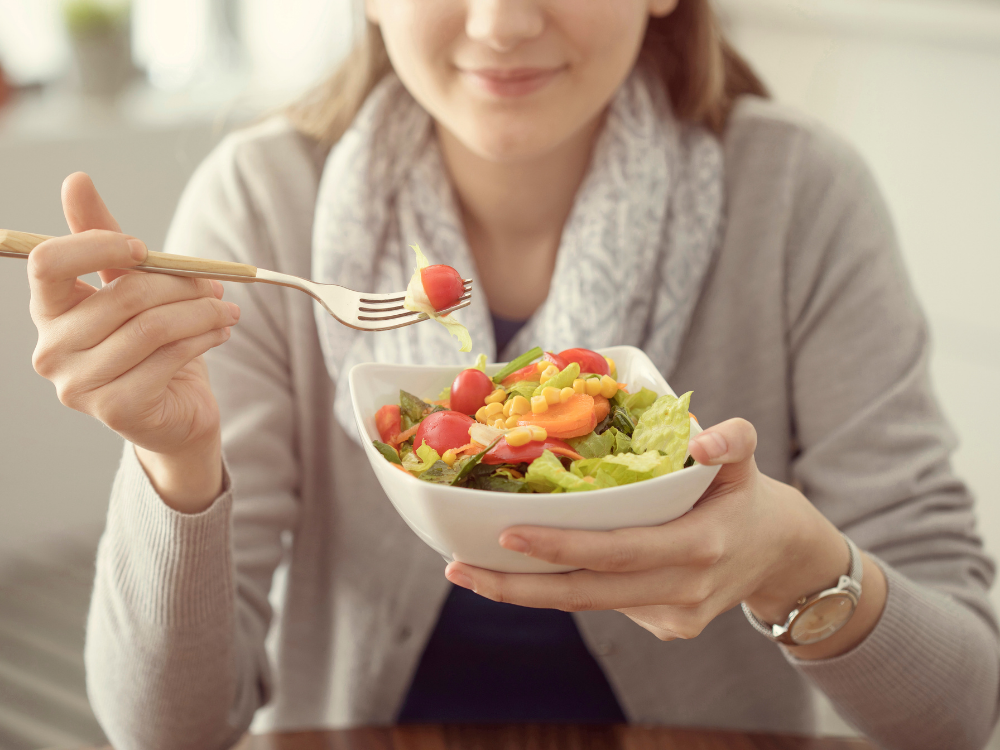 Eine Frau hält eine Schüssel mit Salat, bestehend aus Salatblättern, Kirschtomaten, Mais und Karotten, und isst mit einer Gabel.